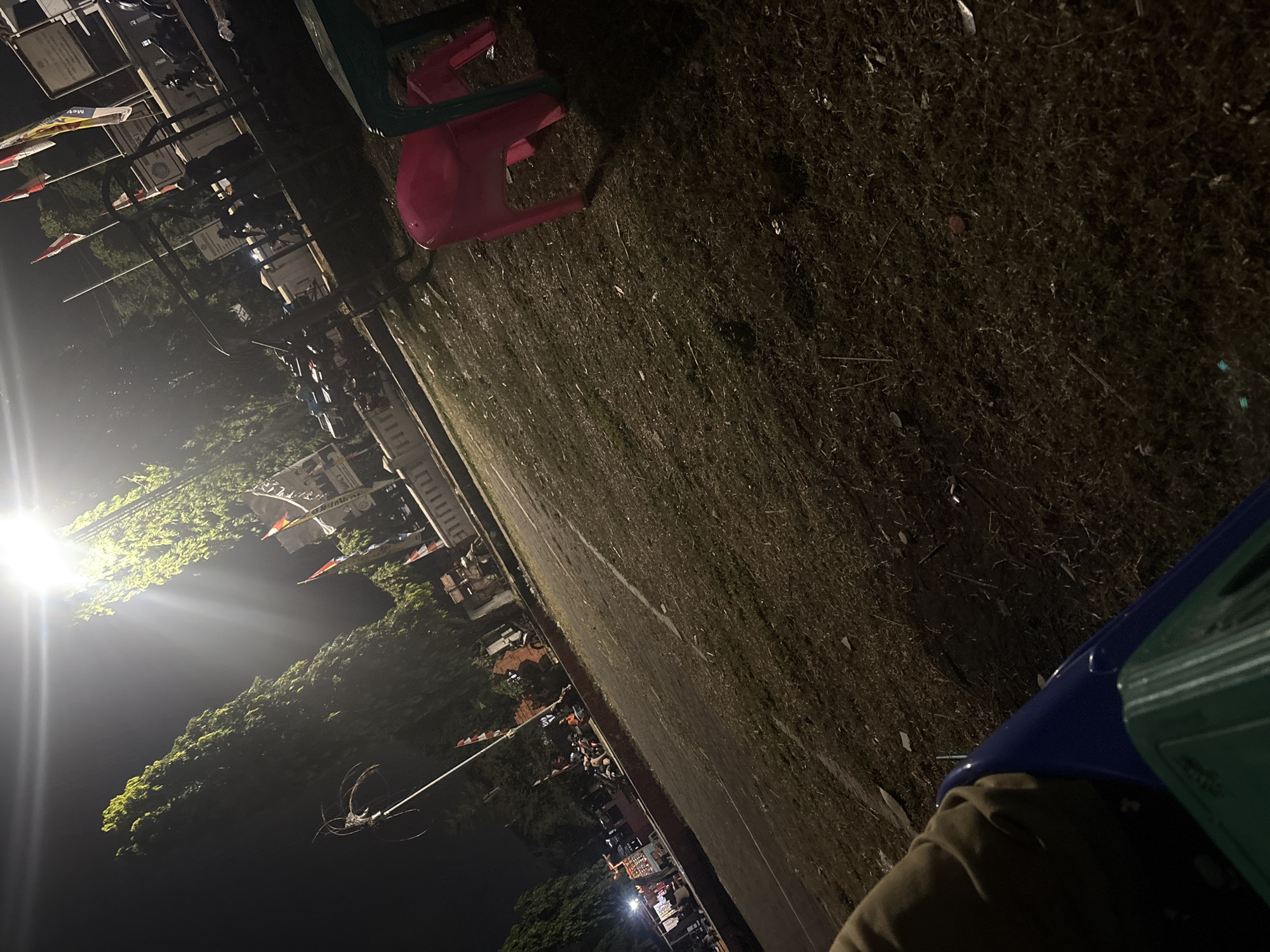 This is a nighttime photograph taken outdoors, likely in Palimanan, Indonesia, as indicated by the presence of multiple flags which might be national or regional. The scene appears to be a public area, possibly a park or a street corner with some vendors. A bright overhead light source illuminates the upper left portion of the image, casting a strong glare and creating lens flare.
In the foreground, a person's arm and shoulder, clad in a brown garment, are visible from a low vantage point, suggesting the photograph was taken from a sitting position or a low camera angle. Partially visible in the lower left is a blue object, possibly a chair or bin, and a green object, which could be another chair or a table.
Dominating the mid-ground is a patch of dry, sparse grass with some debris scattered on it. To the right of the grass, a bright pink plastic child's chair is visible, along with the leg of a green plastic table. Behind the grass and chairs, a paved area is present, with a distinct diagonal line suggesting a pathway or edge.
Further in the background, several trees with dense foliage line a raised concrete structure. This structure appears to be a low wall or platform, possibly part of a building or public monument. On top of this structure and in front of it, various activities are taking place.
Several motorcycles are parked in the background. Some individuals are standing or sitting near these motorcycles and some stalls or small shops. A few flags are displayed on poles, one of which appears to be red and white, possibly the Indonesian flag. One flag has a predominantly yellow and red design. There are also some signs or banners with text on them, though the details are illegible due to the distance and lighting conditions. The overall impression is of a moderately busy, informal public space at night.