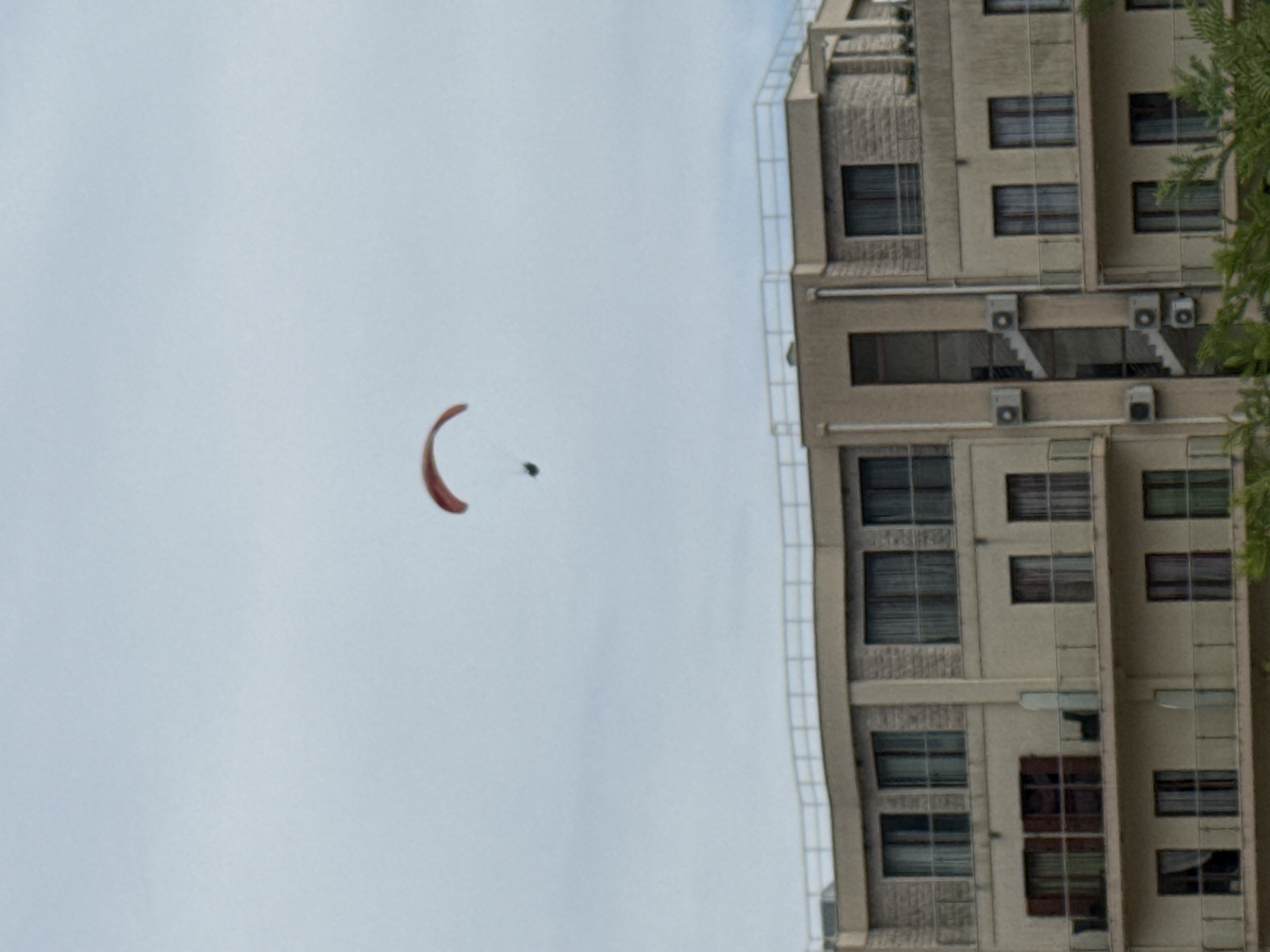In Boreti, Montenegro, a paraglider is visible against a pale, overcast sky, positioned above a multi-story building. The building features multiple balconies with railings and numerous windows, some of which are draped with curtains. The paraglider, with a red canopy, is suspended in the air, and a person is discernible beneath it, appearing to be in mid-flight. The lower portion of the image is partially obscured by the tops of green trees.