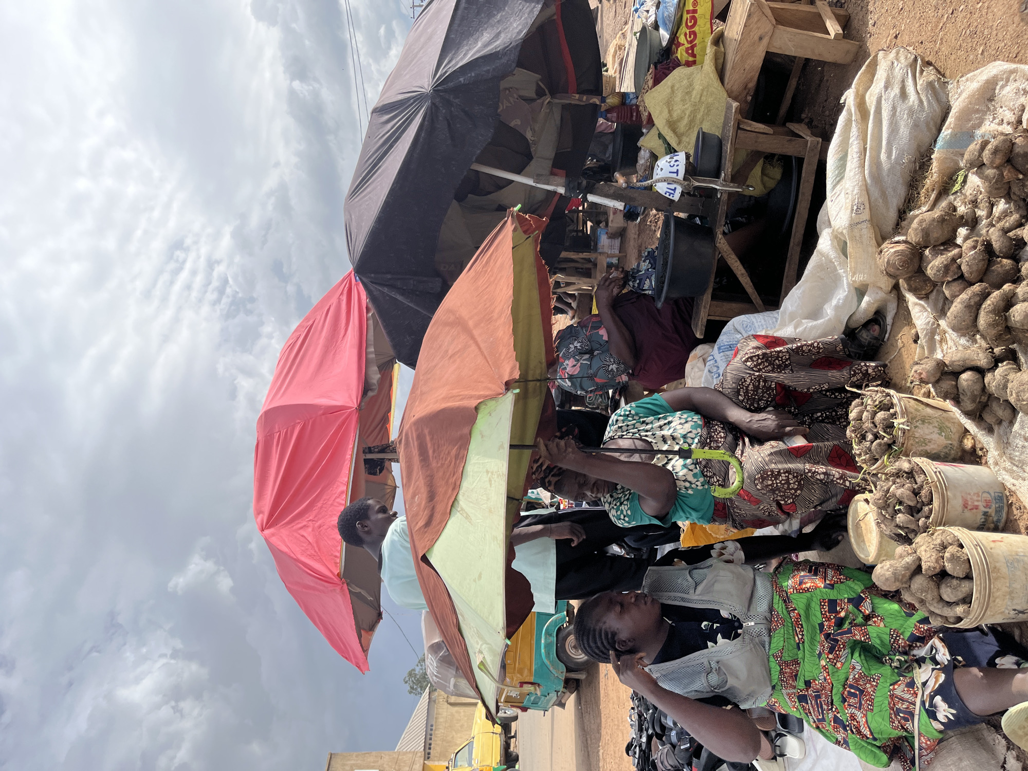 In Rafin Par, Nigeria, under a cloudy sky, a bustling outdoor market scene unfolds. Three large umbrellas, one red, one orange, and one dark, provide shade for vendors and their wares. Two women are seated, engaged in selling produce. The woman on the left, wearing a grey vest over a patterned shirt and a vibrant green and red skirt, gestures with her left hand while looking to her right. In front of her are buckets filled with root vegetables, possibly yams or cassava. Beside her, a second woman in a green and white patterned top and a brown and white patterned skirt holds a small green umbrella. She also has buckets of root vegetables in front of her. To the right, another umbrella shelters a different vendor, with bags of goods and more produce visible. In the background, a yellow autorickshaw is parked on a dusty road, and buildings can be seen in the distance. The overall atmosphere suggests an active marketplace with vendors and customers interacting.