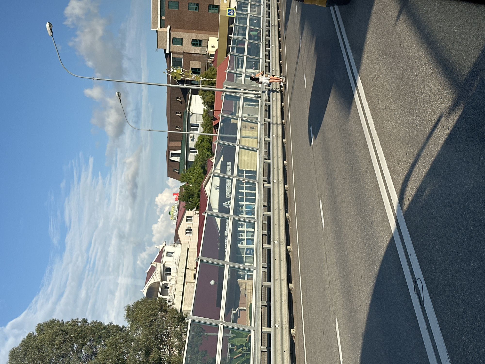 The image depicts a street scene in Sochi, Russia, with a focus on a multi-lane road and the surrounding urban environment. The road, paved with asphalt, is marked with white lane lines. A sound barrier, constructed with glass panels and metal railings, runs along one side of the road. In the background, several buildings with red roofs and a large tree are visible. The sky is blue with scattered white clouds, suggesting fair weather. A person on an electric scooter is seen riding along the road, near the sound barrier. The overall impression is that of a typical day in a coastal city, with elements of modern infrastructure and everyday life.