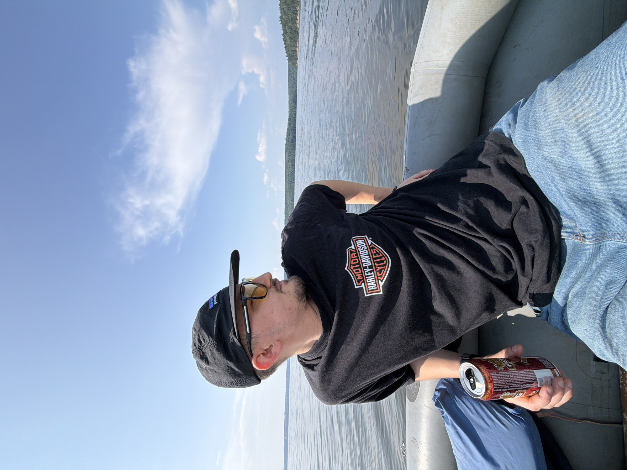 A man wearing a black Patagonia baseball cap, yellow-tinted sunglasses, and a black t-shirt with a Harley-Davidson logo is sitting in an inflatable boat on a body of water. He is holding a red and silver can in his right hand. The boat is moving on a calm lake, with a forest-covered shoreline in the background. The sky is blue with scattered white clouds. The overall scene depicts a leisurely day out on the water. This location is the Krasnoyarskoye reservoir near the city of Divnogorsk, Krasnoyarsk Krai, Russia, not Sylva.