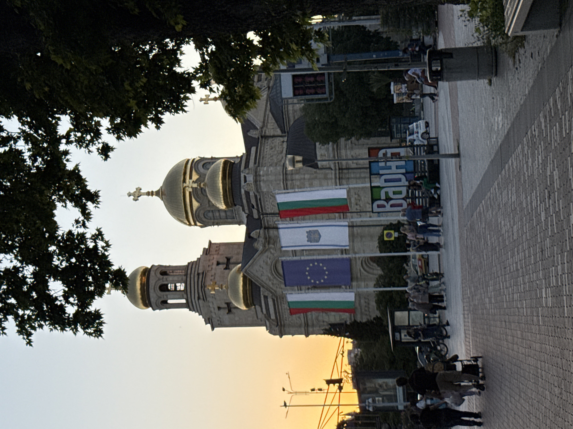 The image shows a wide shot of the Cathedral of the Assumption in Varna, Bulgaria, at sunset. The cathedral's golden domes and cross are visible against the orange sky. In front of the cathedral, there are several flags, including the Bulgarian flag and the European Union flag. A group of people are walking on a cobblestone sidewalk in the foreground, some with shopping bags and a stroller. To the right, a trash can and a bench are visible. In the background, there are streetlights, traffic signals, and buildings. The overall scene is peaceful and serene, with a mix of architectural and natural elements.