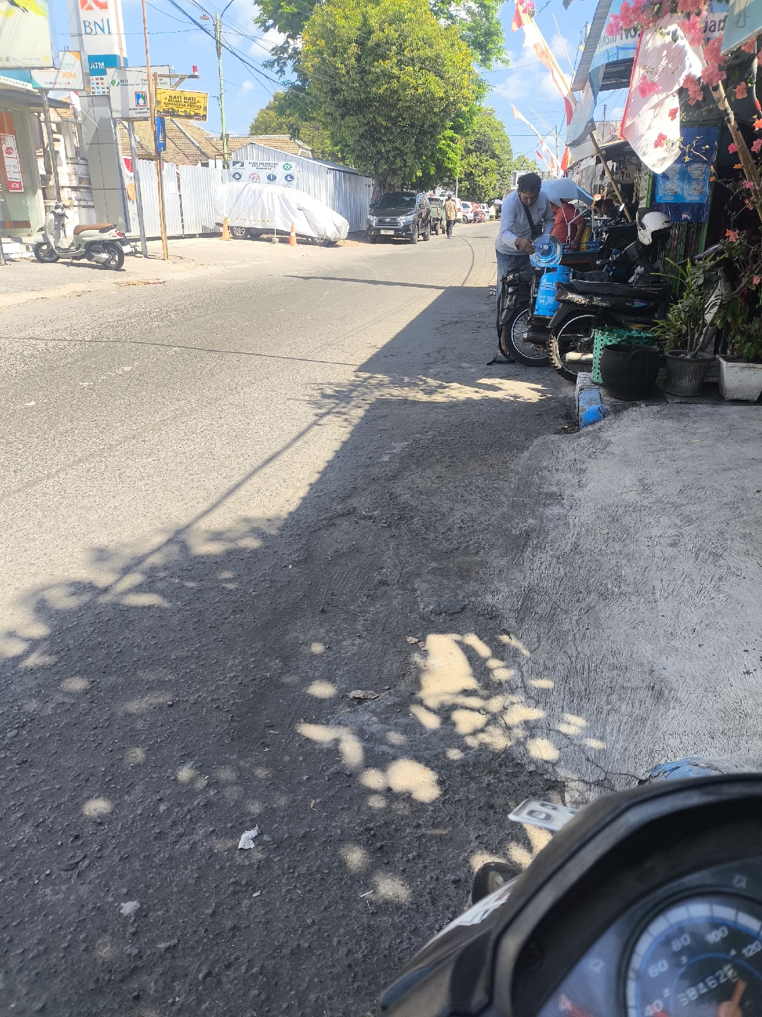 In Lumajang, Indonesia, a man is seen handling a large blue water jug next to a motorcycle parked on a street. He is wearing a light-colored long-sleeved shirt and grey pants. The motorcycle is dark-colored, and a white helmet is visible on its seat. The scene unfolds on a paved street with a concrete sidewalk on the right, lined with potted plants and various items displayed for sale, including posters and flags. Further down the street, construction fencing encloses a site where a white tarp covers a vehicle. A beige scooter is parked on the left side of the street, in front of a building with a "BNI" sign and ATMs. The environment is sunny, casting shadows from trees onto the road.