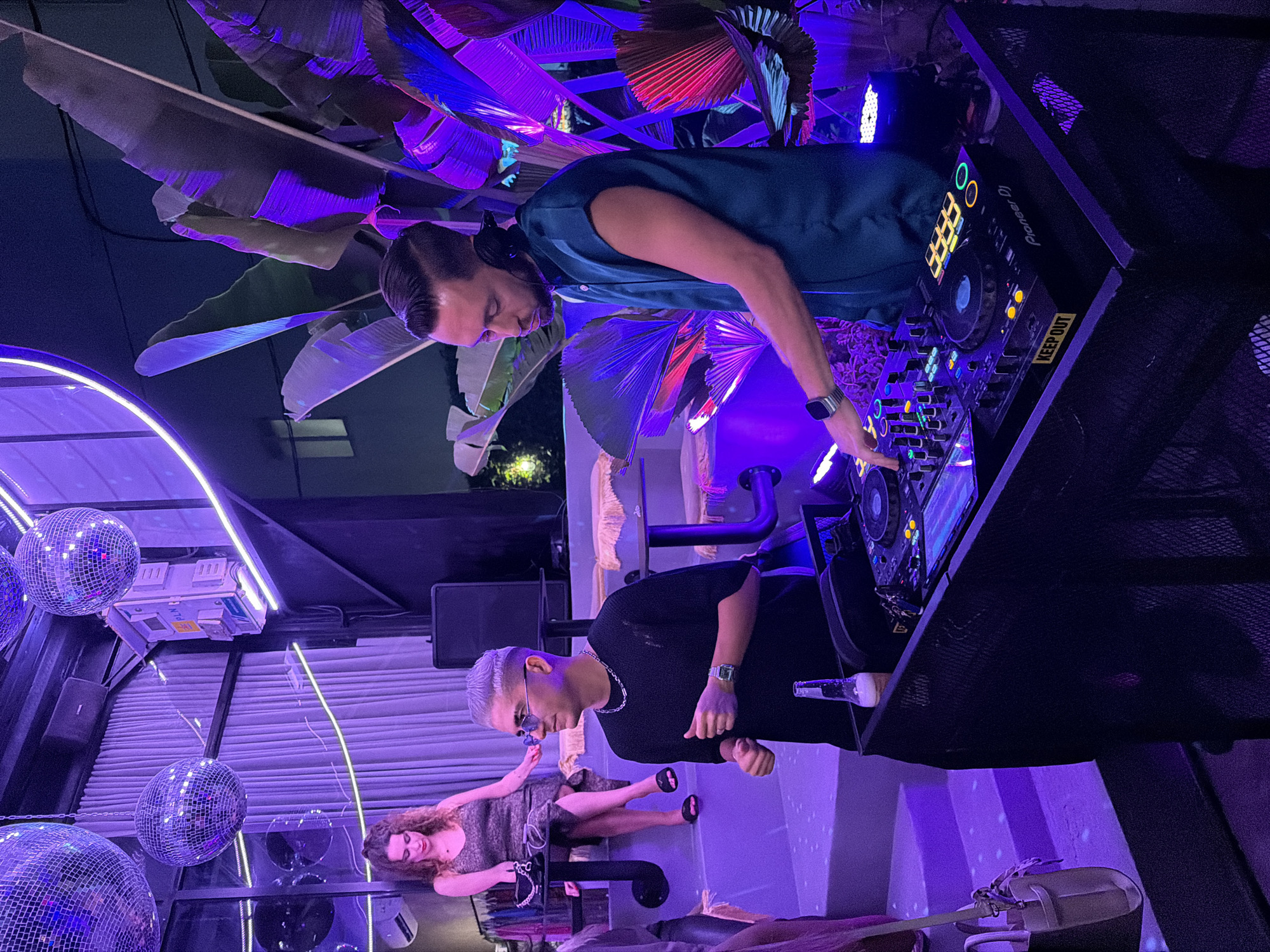 The image captures a vibrant night scene at an outdoor or semi-outdoor venue in North Kuta, Indonesia, likely a bar or club. Two DJs are actively performing, surrounded by tropical-themed decor and dynamic lighting.
In the upper part of the image, one DJ, a man with dark hair, wears a teal sleeveless top and headphones. He is leaning over a Pioneer DJ setup, his right hand adjusting controls, a smartwatch visible on his left wrist. The DJ equipment features glowing buttons, and a yellow "KEEP OUT" sticker is visible on the console. The background is adorned with large, stylized leaves, illuminated in striking purple, blue, and red lights.
In the lower part, another DJ, a man with short, light-colored hair, wearing sunglasses, a black short-sleeved shirt, and a silver chain, stands near another DJ deck. He appears focused, with a watch on his left wrist. Beside him, a clear glass bottle rests on the DJ stand.
To the left, a woman with curly brown hair is seated on a lounge chair or bench, looking down. She wears a patterned dress and sandals. Several mirrored disco balls hang in the foreground and mid-ground, reflecting the surrounding lights. Neon strip lighting traces the architectural elements of the ceiling and walls. The overall atmosphere is lively and energetic, characteristic of a nightlife setting. The venue has a modern design with industrial elements, featuring metal mesh structures. "Pioneer DJ" is visible on the equipment. The time is clearly night due to the dark sky and reliance on artificial lighting.