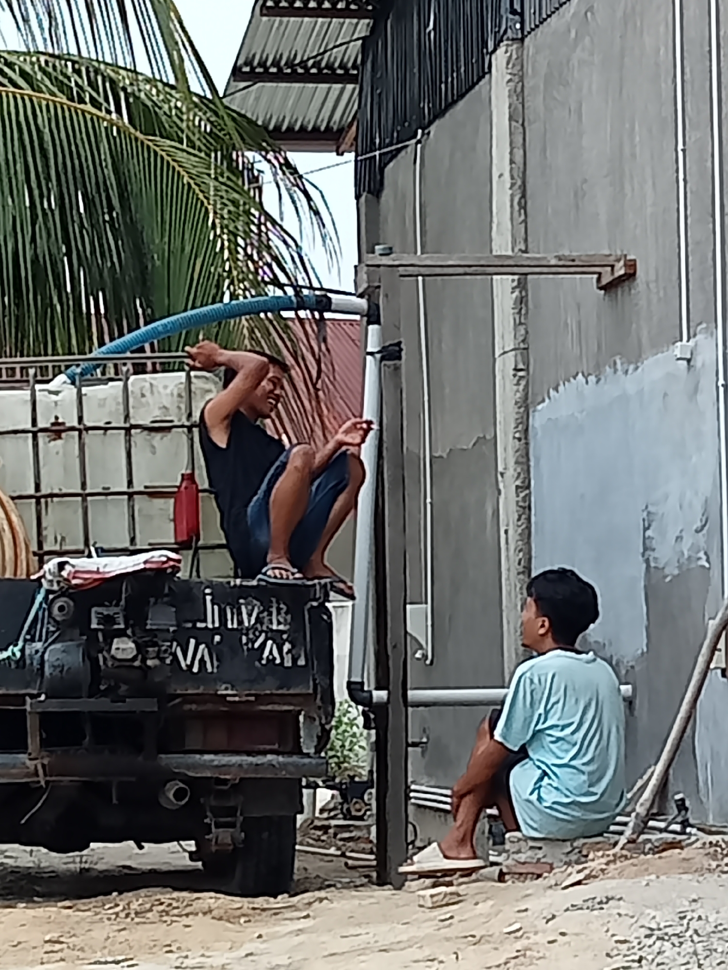 In Melayu Besar Kota, Indonesia, two men are present. One man, wearing a black tank top and blue shorts, is seated on the back of a black truck, holding a blue hose with both hands. He appears to be laughing. The truck is filled with several clear plastic containers. To the right, a second man, wearing a light blue t-shirt and black shorts, is seated on a low brick structure. He is looking away from the truck, towards the right side of the frame. The environment suggests an outdoor setting with a building under construction or repair, indicated by the concrete wall and scaffolding-like structures. Palm fronds are visible in the upper left corner of the image.