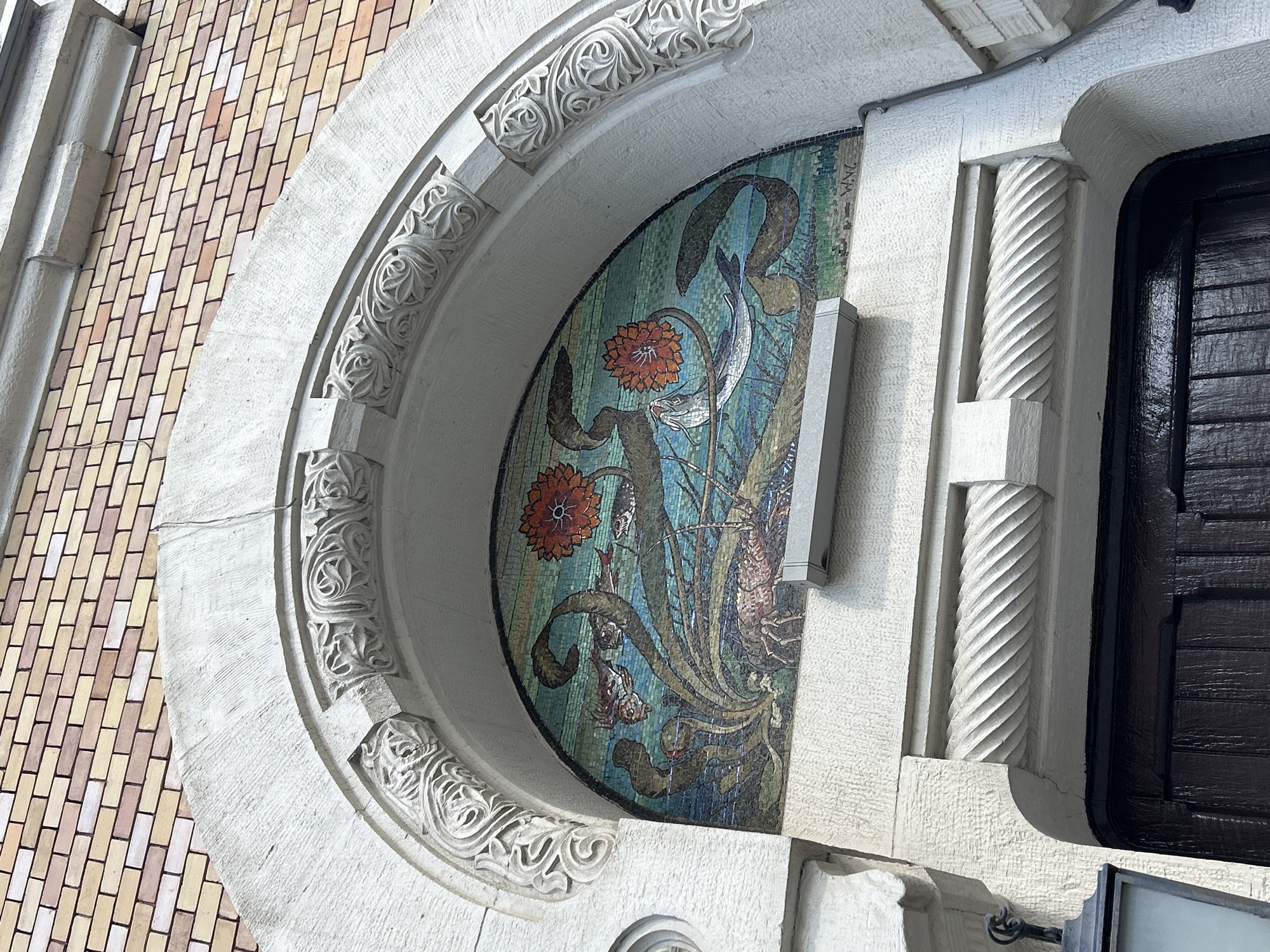 A close-up, eye-level shot showcases a portion of a building's facade in an unknown city. A semi-circular arch frames a mosaic panel, which depicts stylized flowers and leaves, and appears to be a floral mosaic. The arch itself is adorned with intricate carvings, while the surrounding stonework is detailed with twisted pilasters. A dark, possibly wooden door is partially visible to the right of the mosaic. The building’s exterior is a pale color, contrasting with the colorful mosaic and the dark door. The brickwork visible on the left side of the arch is made up of multi-colored bricks.