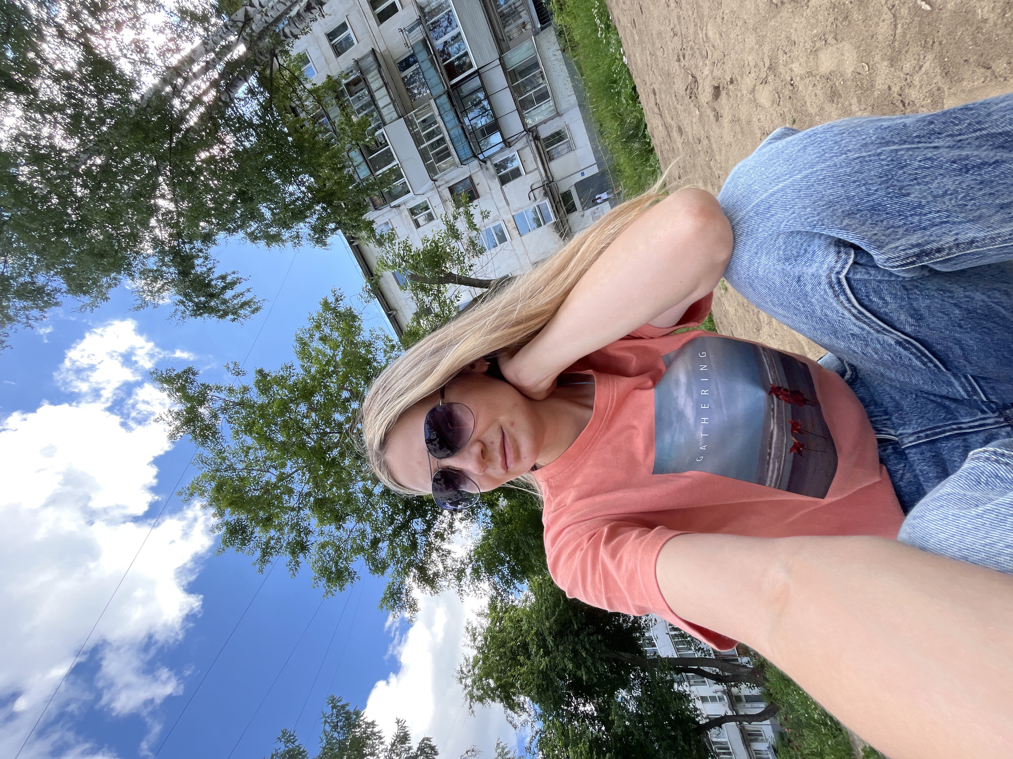 A blonde woman wearing sunglasses, a coral t-shirt with a "GATHERING" graphic, and blue jeans is sitting on sand. She is resting her head on her hand and looking to her left. Behind her, there is a multi-story apartment building, green trees, and a blue sky with white clouds. The image appears to be taken in the city of Chaikovsky, Russia.