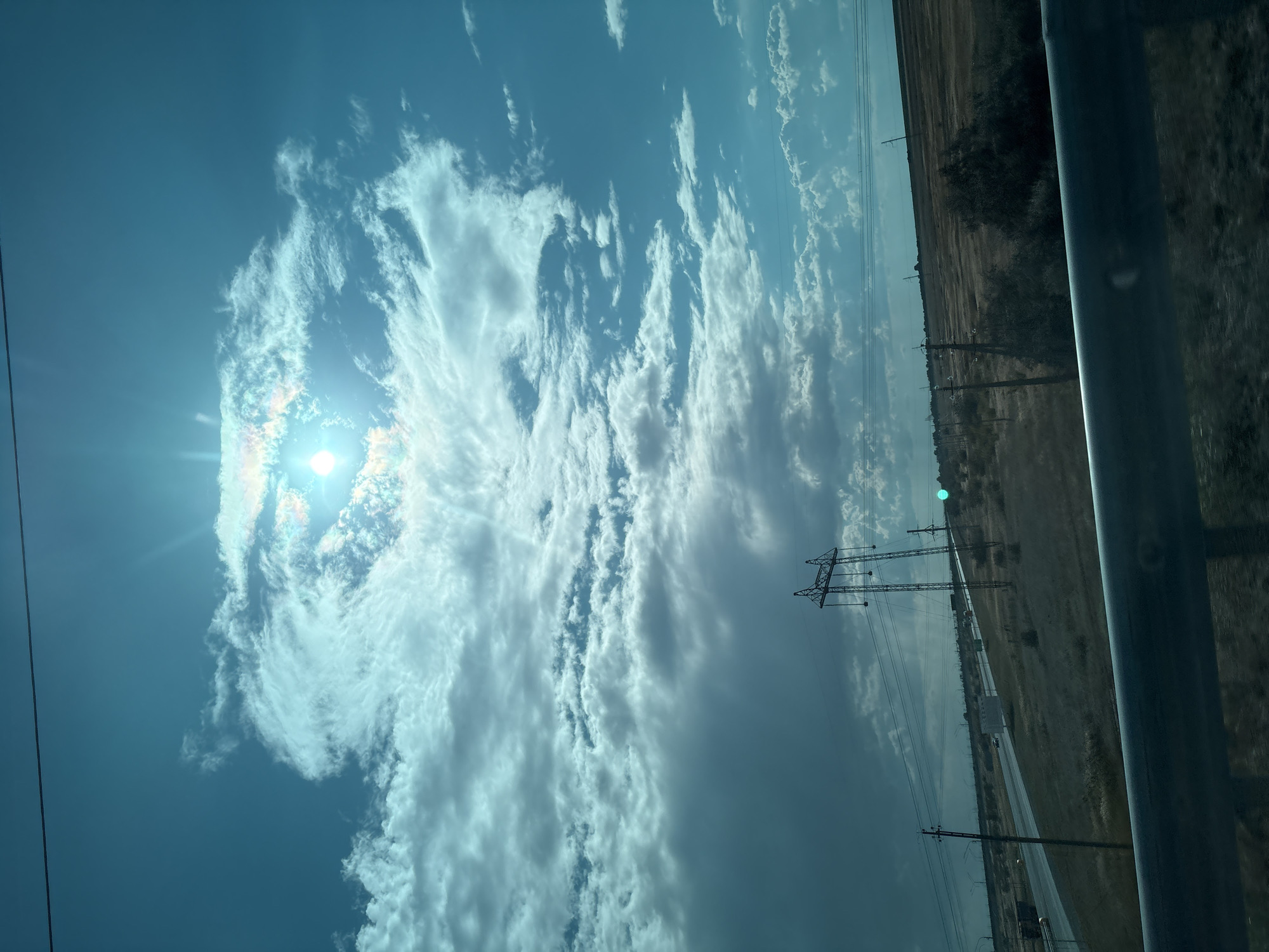 The image captures a wide-angle view of a vast, open landscape under a dramatic sky. The sky is a prominent feature, filled with an array of clouds ranging from wispy cirrus formations near the sun to darker, more ominous cumulus clouds in the distance. The sun, partially obscured by clouds, shines brightly, creating a lens flare effect and casting light rays.
The landscape below consists of dry, golden-brown grassland and scattered bushes. A dark, paved road stretches across the middle ground, with several vehicles visible, suggesting it's a highway. Power lines and utility poles are scattered throughout the scene, some tall and industrial-looking, others smaller and more traditional. The overall impression is one of a rural or semi-rural environment.
A thick, dark metal railing is visible in the foreground, suggesting the photo was taken from a vehicle or elevated position. The color palette is dominated by blues and greens in the sky, contrasted with the browns and grays of the land and infrastructure. There are no people clearly visible in the image. The scene appears to be located in a location with infrastructure such as roads and power lines, typical of an inhabited or traversed region within Kazakhstan, potentially near Атамекен кенттік әкімдігі given the context.