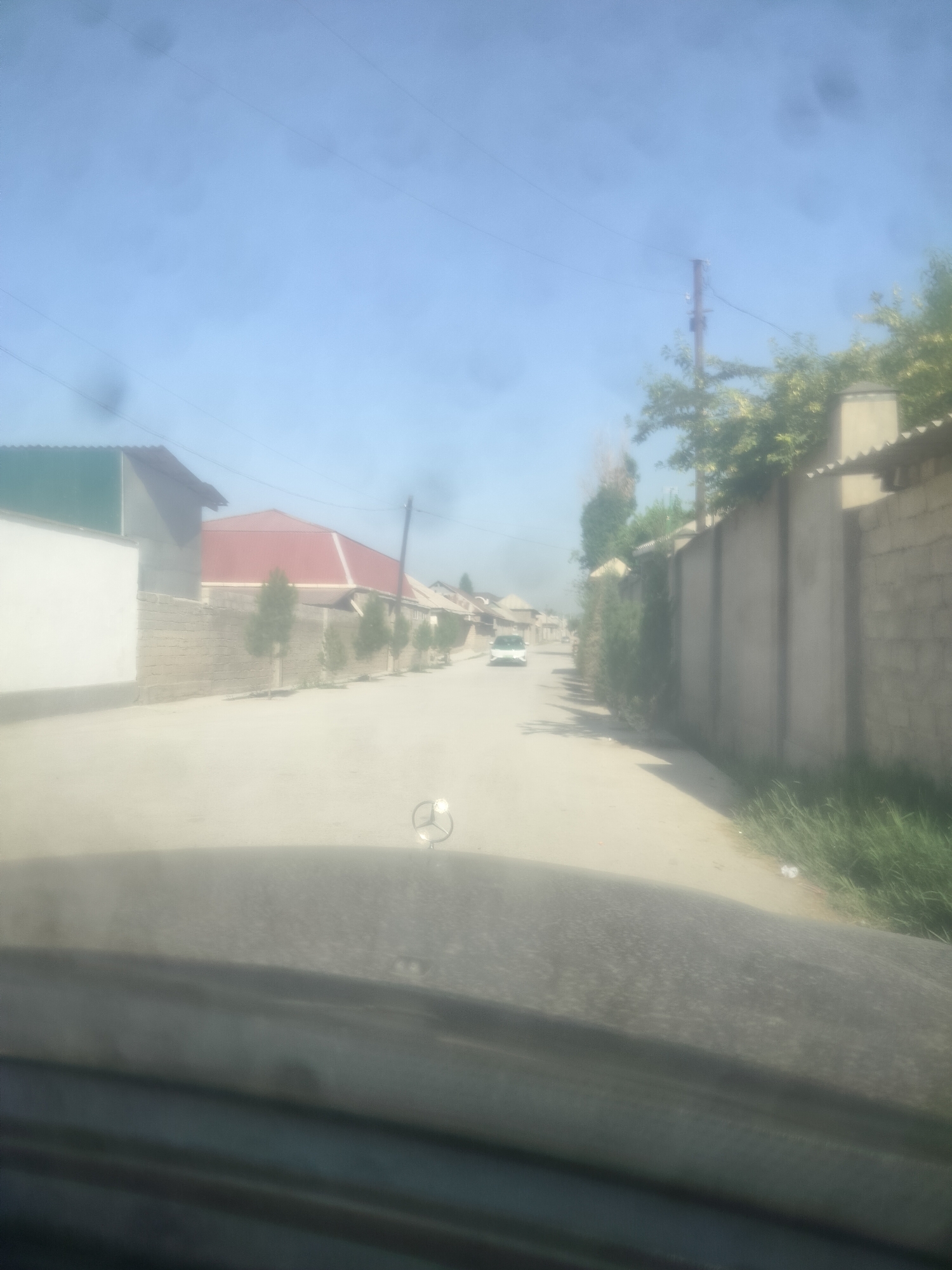 A perspective shot from inside a vehicle looking out onto a street in Navobod, Tajikistan. The street is paved and appears to be lined with residential buildings and walls. On the left side of the street, there is a white wall, a building with a green roof, and a building with a red roof. Several small, conical trees are planted along a brick wall that separates the street from the properties. On the right side of the street, there is a tall, grey concrete wall with a peaked top, and beyond it, trees and vegetation. Further down the street, a white car is visible, and the road curves to the right. The sky is blue with some hazy elements, and a utility pole with wires is visible in the distance. The dashboard of the vehicle is visible in the foreground, with a faint reflection of the Mercedes-Benz logo on the hood ornament. The overall impression is a quiet, residential street in a sunny, possibly warm climate.