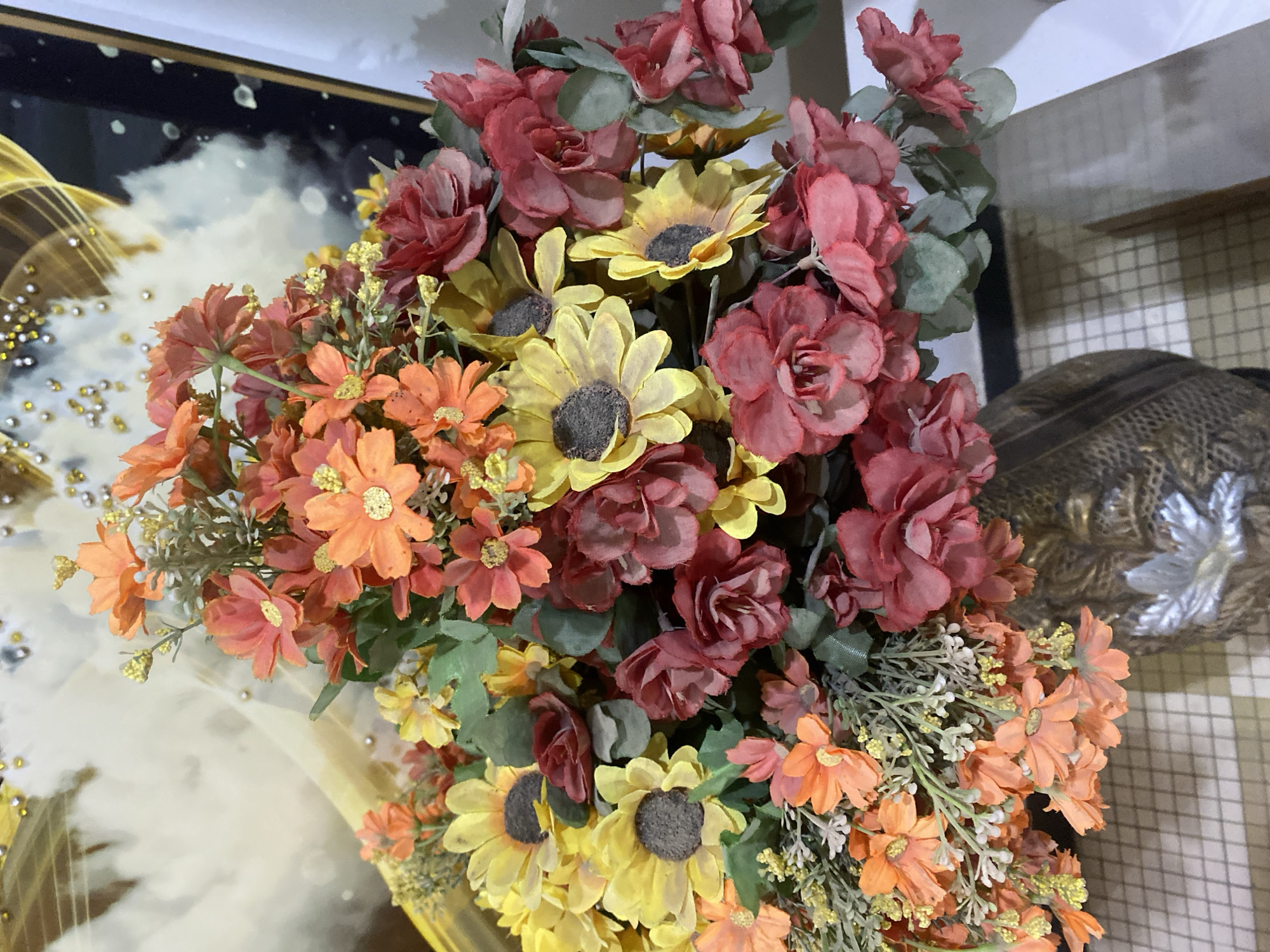 This is a close-up, eye-level, vertical, medium shot of a vibrant artificial flower arrangement in a decorative vase. The bouquet is a mixture of sunflowers, roses, and smaller daisy-like flowers in shades of yellow, orange, and red, interspersed with green foliage. The vase itself appears to be made of a textured, metallic material with an ornate pattern. In the background, a framed piece of art with abstract golden and white swirls is visible, along with a white wall. The flowers are the primary focus, filling most of the frame. There are no people or animals present, and no actions are taking place. The scene is indoors, likely in a home or decorative setting. The information about Giginyu, Nigeria is not supported by the visual content of the image.