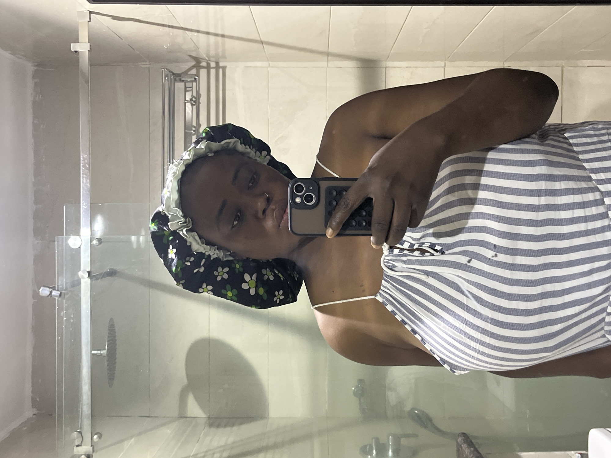 A woman is taking a selfie in a bathroom. She is wearing a floral shower cap and a striped pajama top. The bathroom has white tiled walls, a glass shower door, and chrome fixtures. The location is a bathroom, no information to indicate the city of Abuja, Nigeria.