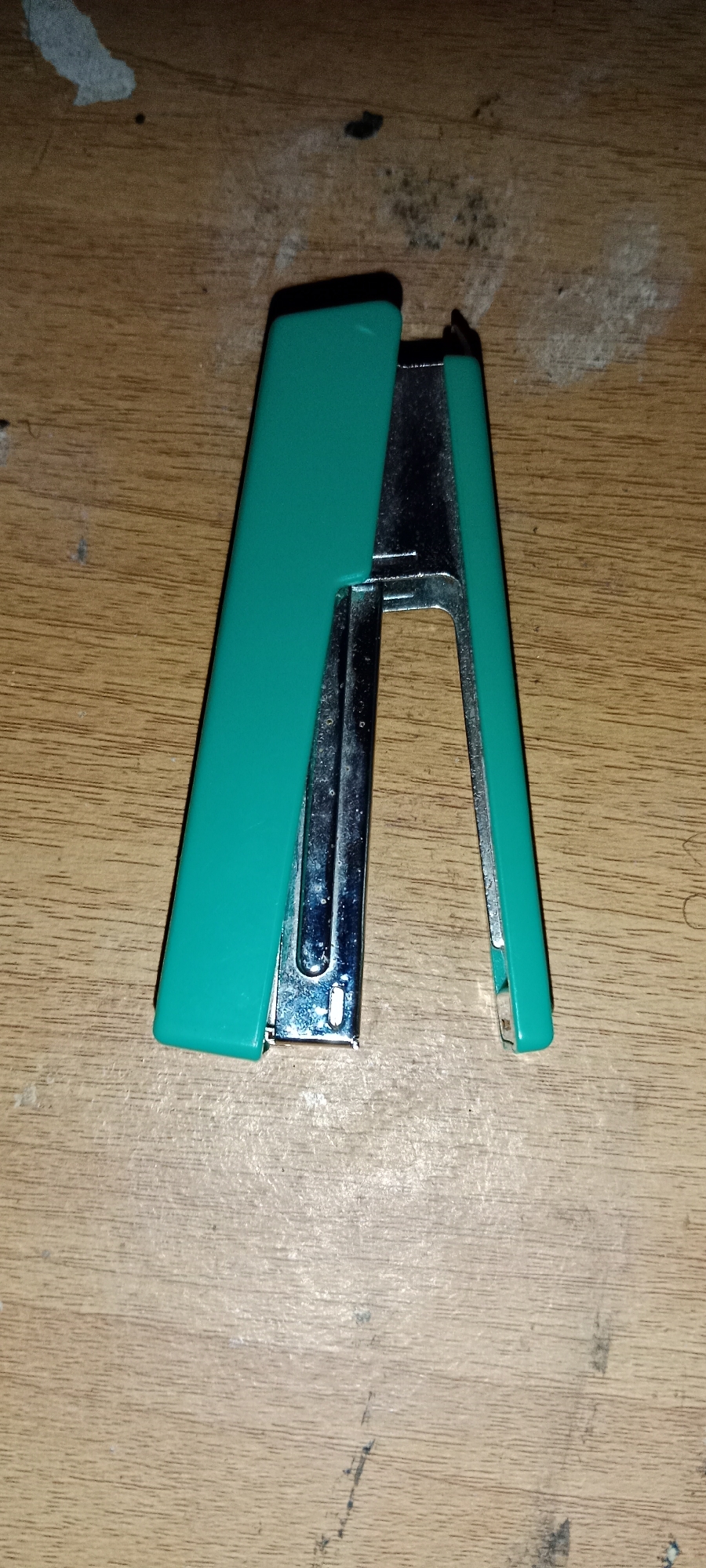 A teal-colored stapler is centered on a wooden surface. The stapler is open and angled, with its two halves spread apart. The metal components of the stapler are visible, as is the teal plastic casing. The wooden surface appears to be a table or desk, with a grainy texture and some faint marks or scratches. The lighting is overhead, casting a shadow beneath the stapler. There are no people or other objects visible in the image. The scene is indoors, likely a home or office environment.
