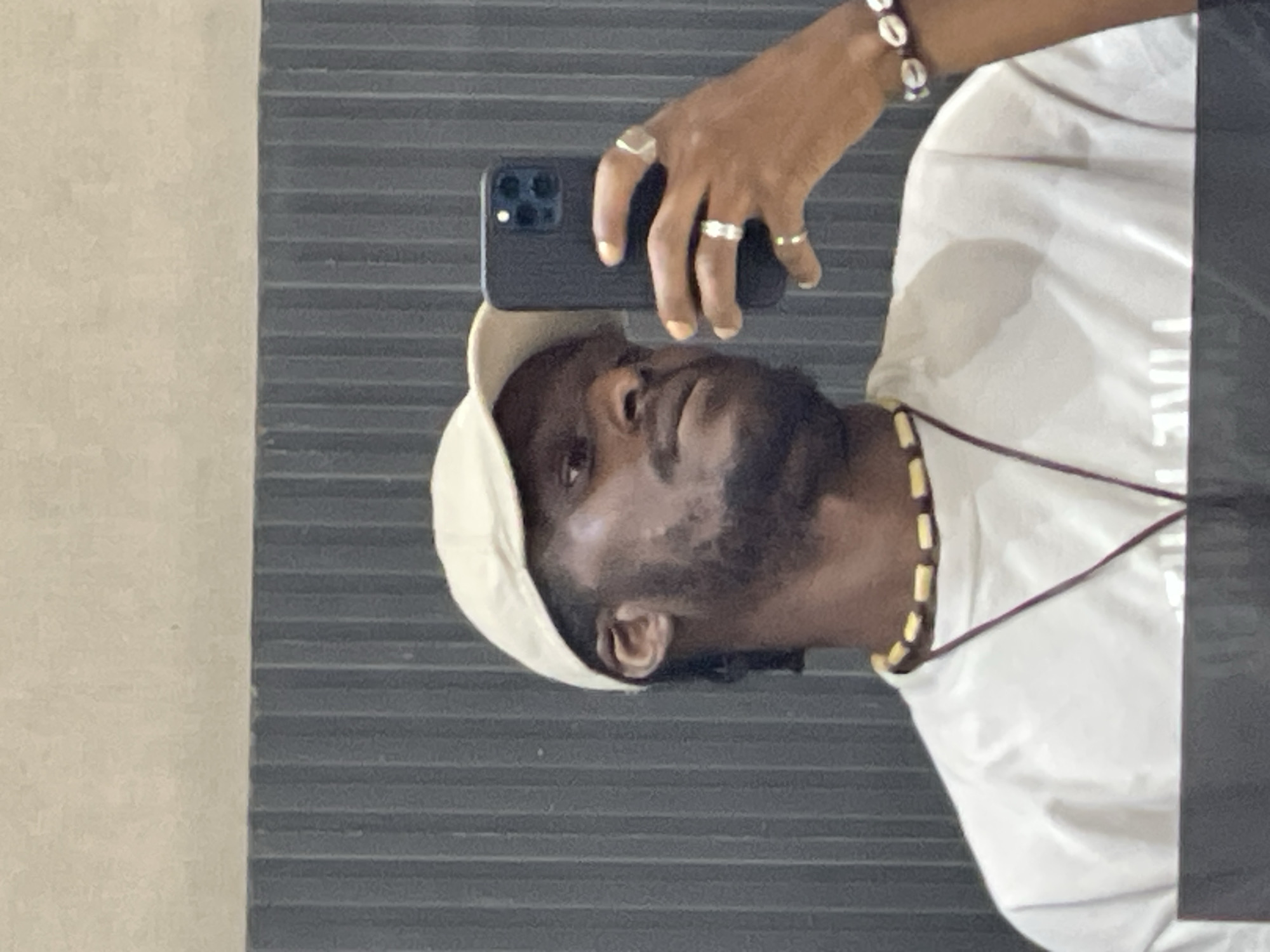 A selfie of a Black man wearing a light-colored baseball cap and a white t-shirt is reflected in a mirror. He has a beard and is wearing a necklace with wooden beads and a bracelet made of seashells. He is holding a black smartphone in his right hand, which is raised to take the photo. The background consists of a ribbed, dark gray wall and a lighter-colored wall above it. The image is taken indoors, likely in a bathroom or changing room. It is not possible to determine the exact location in Guzape, Nigeria from the image alone.