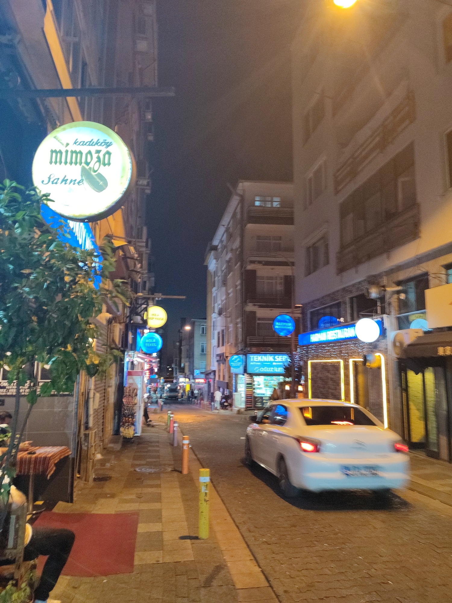 This is a night-time, eye-level, medium shot of a street scene in Kadıköy, Turkey. On the left side of the street, a round, illuminated sign with "kadıköy mimoza Sahne" written on it hangs above a green tree. The street is paved with cobblestones and there is a sidewalk with yellow and orange bollards. A white car is driving away from the viewer down the street. Buildings line both sides of the street, with various signs and lights visible. The sky is dark and hazy.
