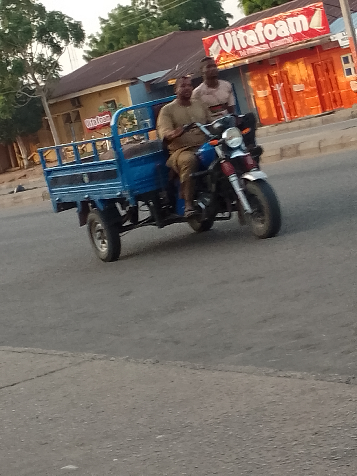 In Pantame, Nigeria, two individuals are riding a blue three-wheeled motorcycle. The motorcycle has a cargo bed at the rear. The driver, wearing a light brown shirt and khaki pants, is operating the vehicle. A passenger, wearing a patterned shirt, is seated behind the driver. They are on a paved road, with buildings and trees visible in the background. A red and white sign with the word "Vitafoam" is prominently displayed on one of the buildings. The scene appears to be during daylight hours, possibly late afternoon due to the lighting.