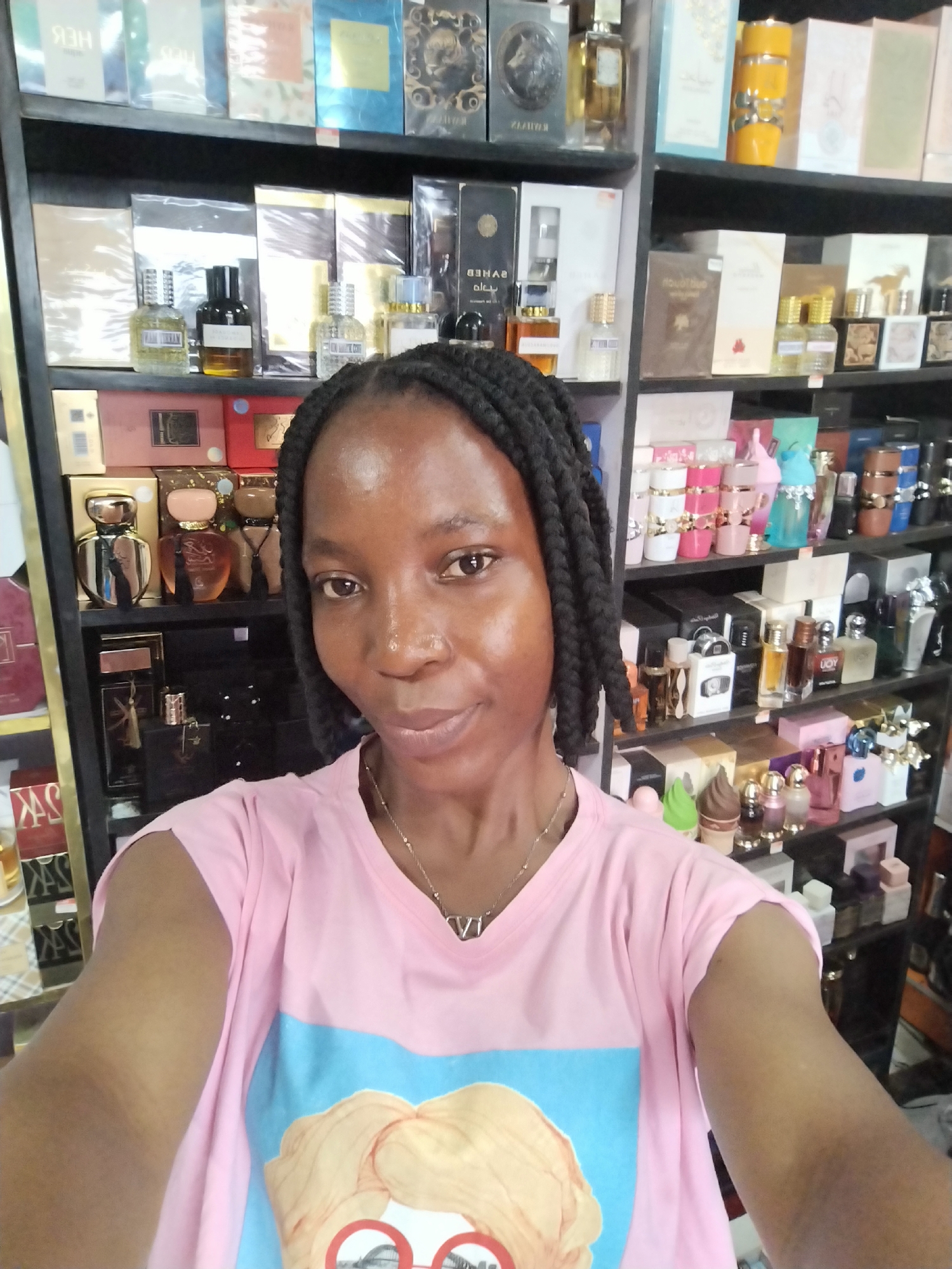 A young Black woman with braided hair is taking a selfie in front of shelves stocked with perfumes. She is wearing a light pink t-shirt with a light blue and yellow graphic design on it, and a delicate silver necklace. The background consists of multiple shelves filled with various perfume bottles and boxes, suggesting she is in a retail environment, possibly a perfume shop or stall. The lighting is consistent with indoor retail lighting. The image appears to have been taken in Ugbowo, Nigeria, based on the provided information.