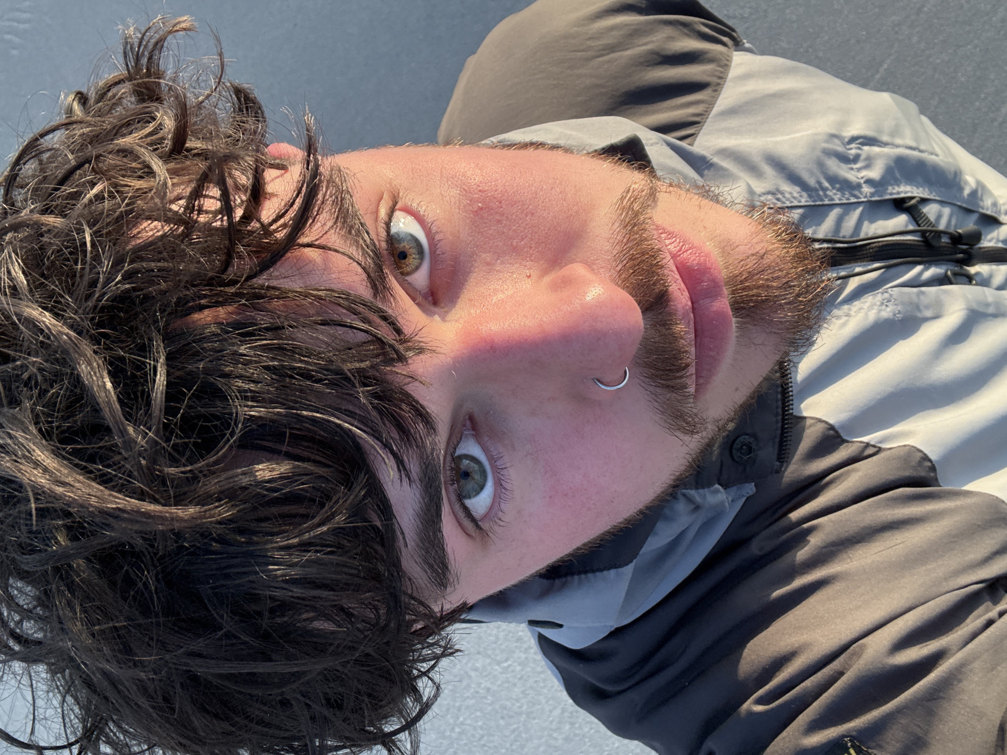 A young adult male is centered in the image, gazing directly at the camera with slightly wide eyes, suggesting surprise or alertness. He has hazel-green eyes, fair skin, dark brown curly hair, and a short, scruffy beard and mustache. A silver hoop earring is visible in his left nostril. He's wearing a dark gray and light gray jacket with a zippered front.
The background appears to be a light gray, possibly concrete or asphalt. The lighting is bright and seems natural. There's no discernible city location indicated. Therefore the location is Unknown, Unknown.
