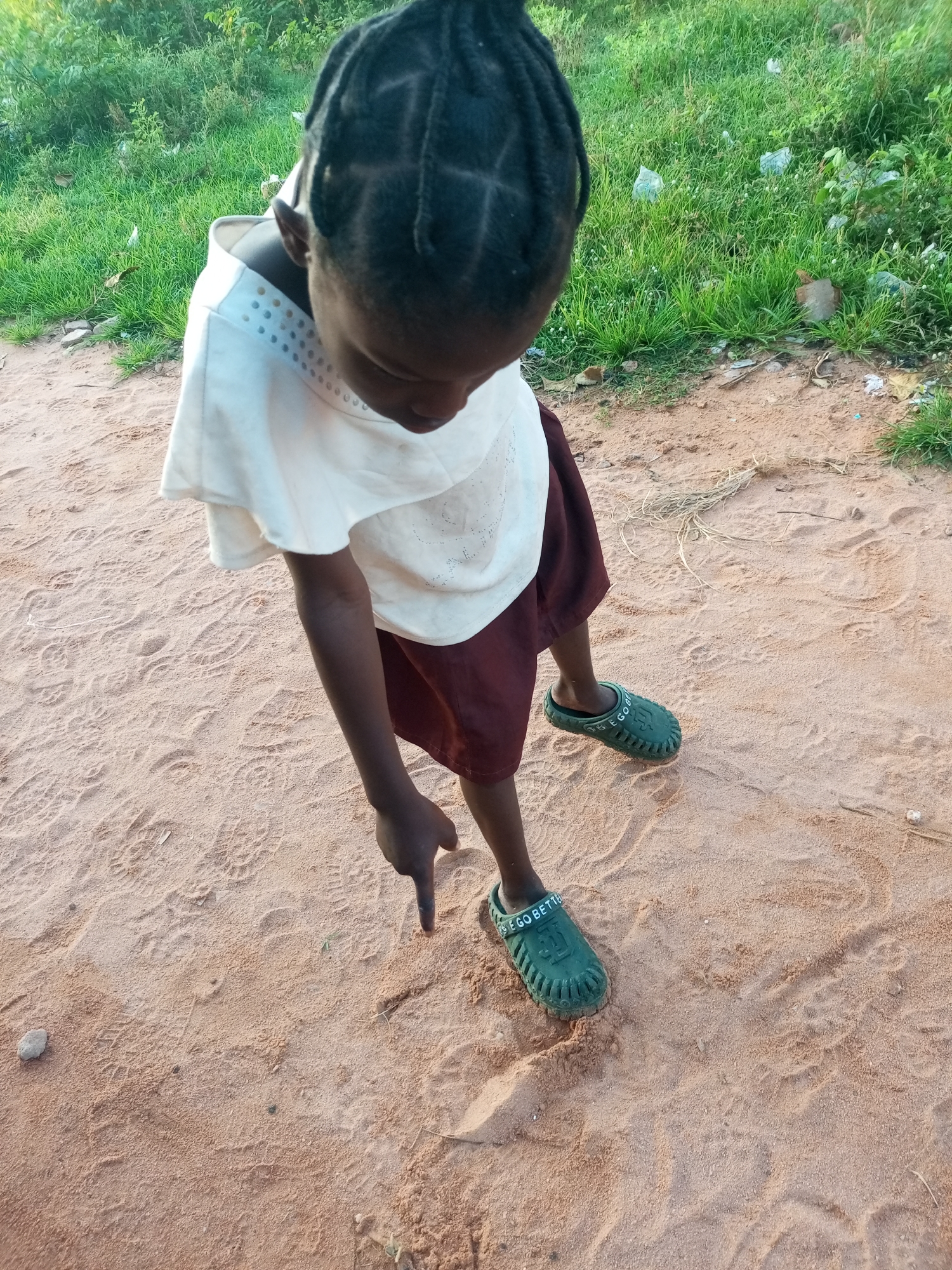 A young Black child, facing downward, points to the sandy ground with their right index finger. The child wears a white shirt with a decorative neckline and a maroon skirt. On their left foot, they wear a green clog with the inscription "GO BETTE" visible. The ground is composed of reddish-brown sand, with some visible footprints and debris. To the upper right of the frame, green grass and vegetation can be seen. The scene appears to be outdoors in a rural or semi-rural area, possibly in Tempure, Nigeria, given the vegetation and sandy soil.