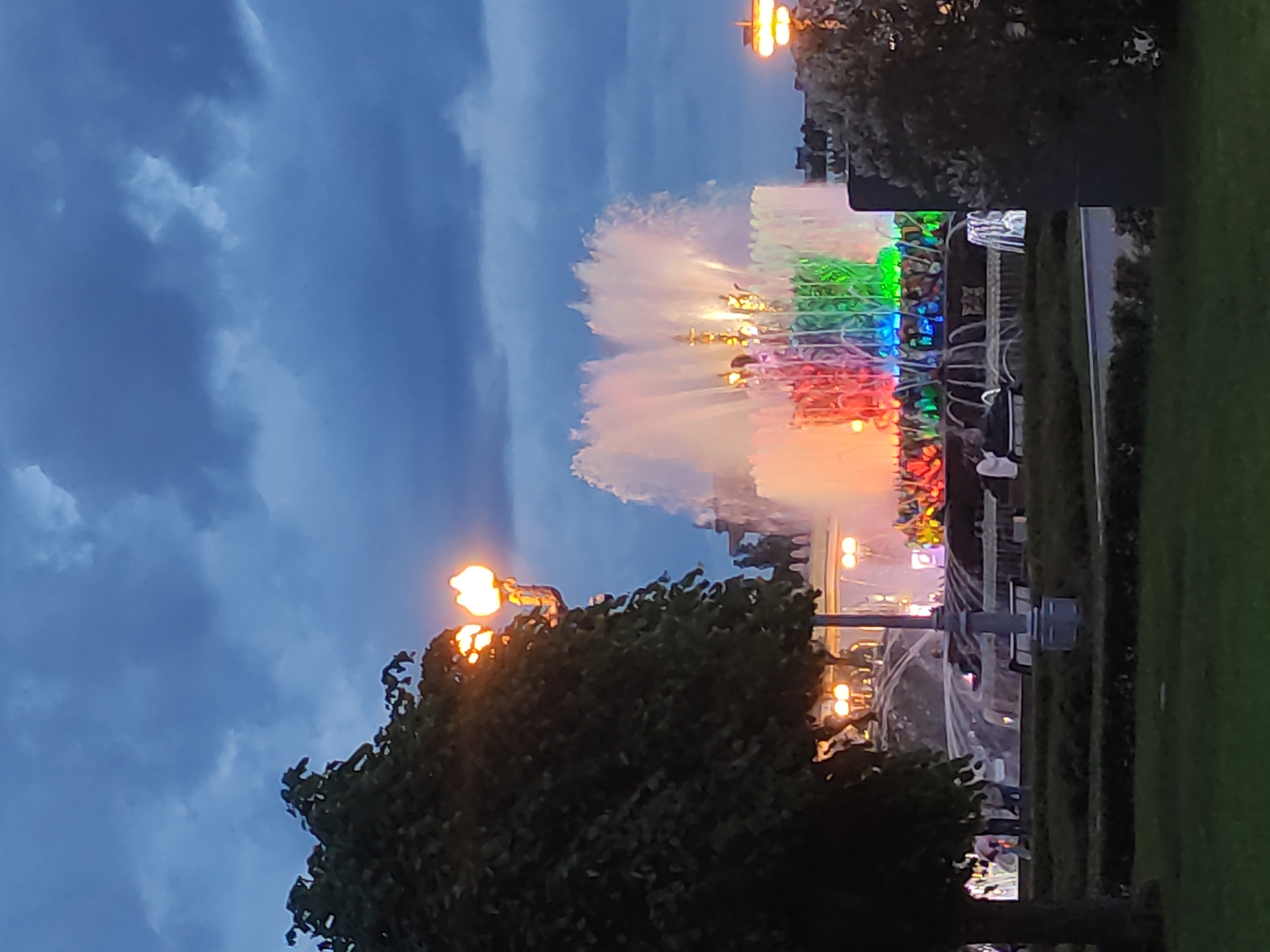 The image depicts a water fountain illuminated with a spectrum of colors, ranging from red to green, located in Moscow, Russia. The fountain is prominently featured, with water jets reaching upwards and reflecting the colored lights. The sky in the background is a deep blue, suggesting either dusk or early evening. Two dark green trees frame the bottom portion of the image, and street lights with orange bulbs are visible near the trees and the fountain. People are present near the fountain, some appearing to watch the spectacle. A patch of green grass lines the right side of the frame.