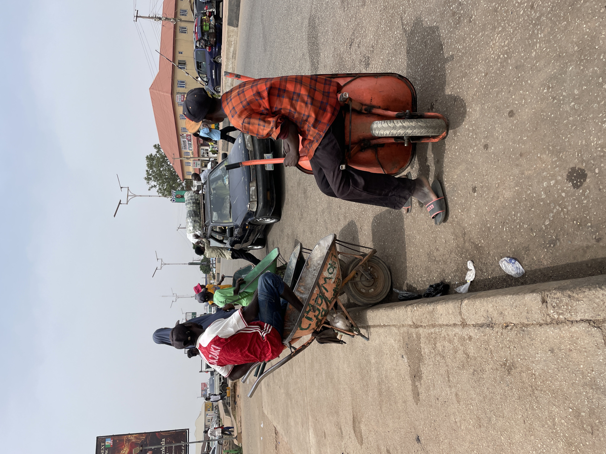 In Jos, Nigeria, the image shows two individuals sitting in wheelbarrows. The person in the foreground wears a red and white jersey. A man in the background wears a red and orange flannel shirt and pushes his wheelbarrow. Cars and other people are visible in the background, along with buildings and streetlights. There is litter scattered on the curb.