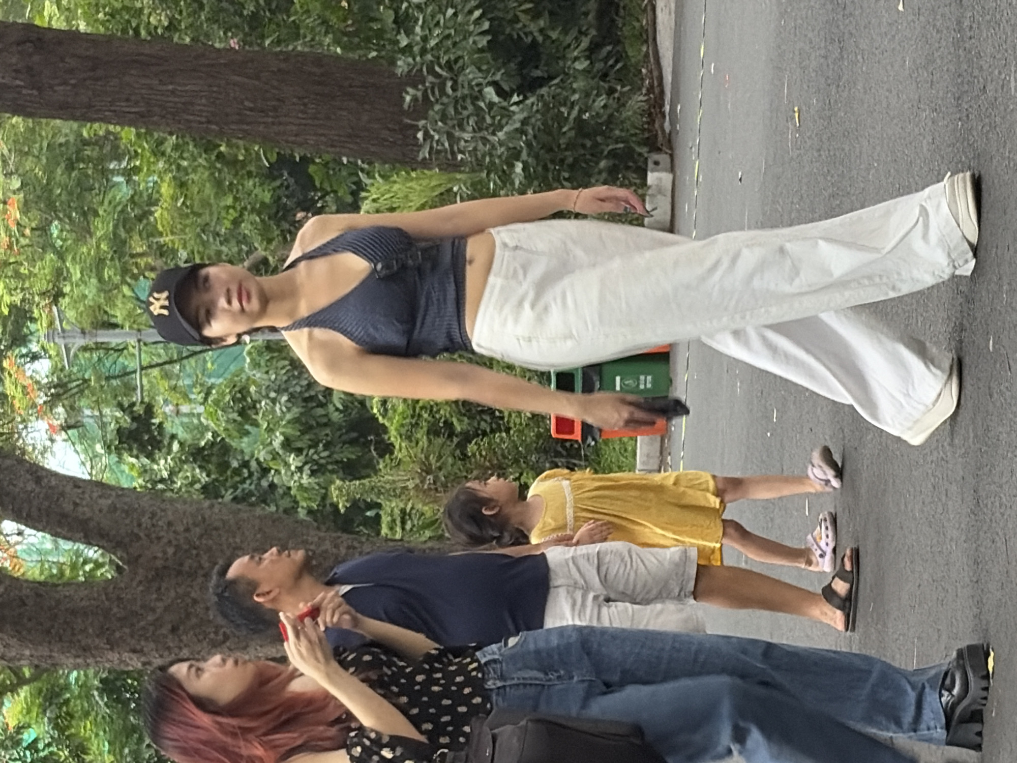 This image captures an outdoor daytime scene in Thủ Đức, Vietnam, depicting four individuals walking on a paved pathway. The background features dense green foliage, mature trees with prominent trunks, and hints of red-orange blossoms.
From left to right:
A young adult female with reddish-brown hair, wearing a black and white off-shoulder polka dot top, blue jeans, and black chunky platform shoes, holds a red object in her hands. A black bag is slung over her left shoulder.
Beside her, an adult male with short dark hair wears a navy blue polo shirt, light-colored shorts, and black slide sandals. He is walking while holding the right hand of a young girl.
The young girl, with dark hair tied back, wears a yellow short-sleeved dress with white trim and light-colored (possibly pink or purple) crocs.
On the far right, a young adult female wears a black New York Yankees baseball cap, a dark blue halter-neck ribbed crop top revealing her midriff, white wide-leg trousers, and white sneakers. She holds a black smartphone in her right hand and has sunglasses tucked into her top and a thin red bracelet on her left wrist.
All individuals are in motion, generally walking from left to right across the frame. The paved ground is dark grey with scattered light-colored debris. In the mid-ground on the right, an orange and a green trash bin with white lettering are visible. A grey pole stands in the left background.