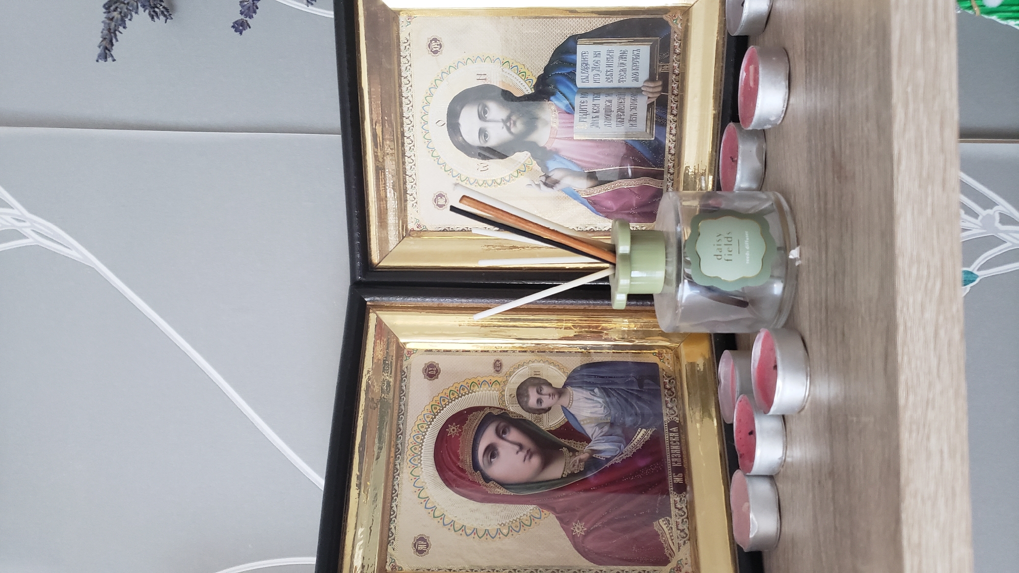 An indoor shot captures a collection of religious icons, tealight candles, and a reed diffuser displayed on a wooden shelf. The two icons, framed in gold, are positioned side-by-side. The icon on the left depicts the Virgin Mary holding the Christ child. The icon on the right portrays Jesus Christ holding an open book. In front of the icons, a clear glass reed diffuser with a light green label that reads "daisy fields" and "reed diffuser" is visible, with several reeds protruding from its top. Surrounding the diffuser are several unlit tealight candles, some with pink wax. The background consists of a light grey wall with a subtle geometric pattern. The scene is lit from above, creating soft shadows. There is no indication of the specific location in Bratislava, Slovakia.