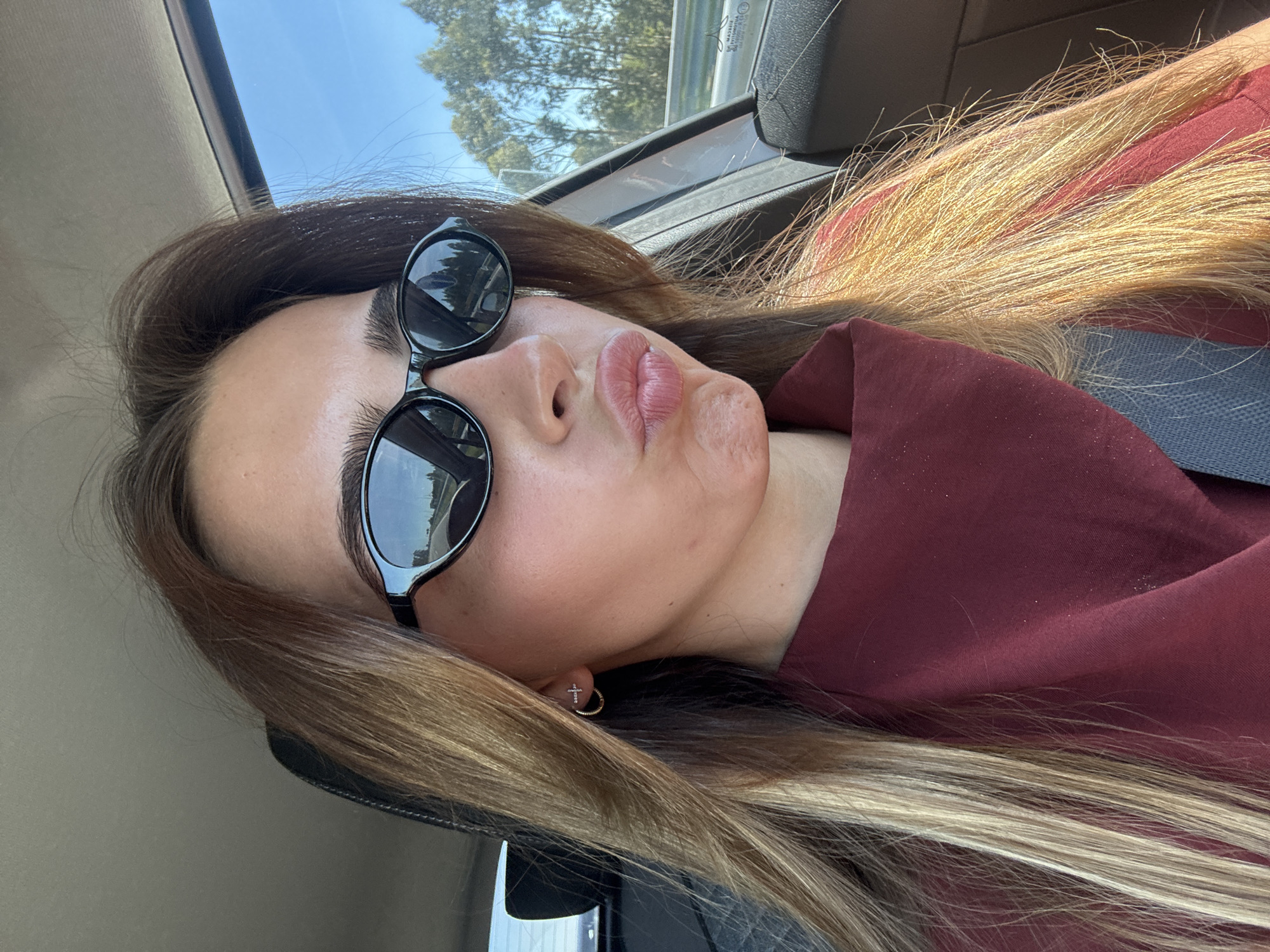 A female subject is depicted in a medium close-up selfie inside a vehicle, likely on a road within the Aveiro, Portugal region. She has long, multi-toned brown hair with prominent blonde highlights. Her face is directed towards the camera, with lips pursed in a "kiss" pose. She wears black, oval-shaped sunglasses that obscure her eyes. Two small, gold-colored earrings are visible on her right ear: a small hoop and a cross-shaped stud. Her attire consists of a dark red garment, possibly a top, with visible folds around the neck area. A portion of a blue fabric is visible below the red garment on the lower left. The vehicle's interior features a light gray headliner and a black headrest. Outside the car, visible through the right window and reflected in her sunglasses, is a clear blue sky, a road with a guardrail, and trees with light-colored trunks, consistent with a sunny daytime environment.