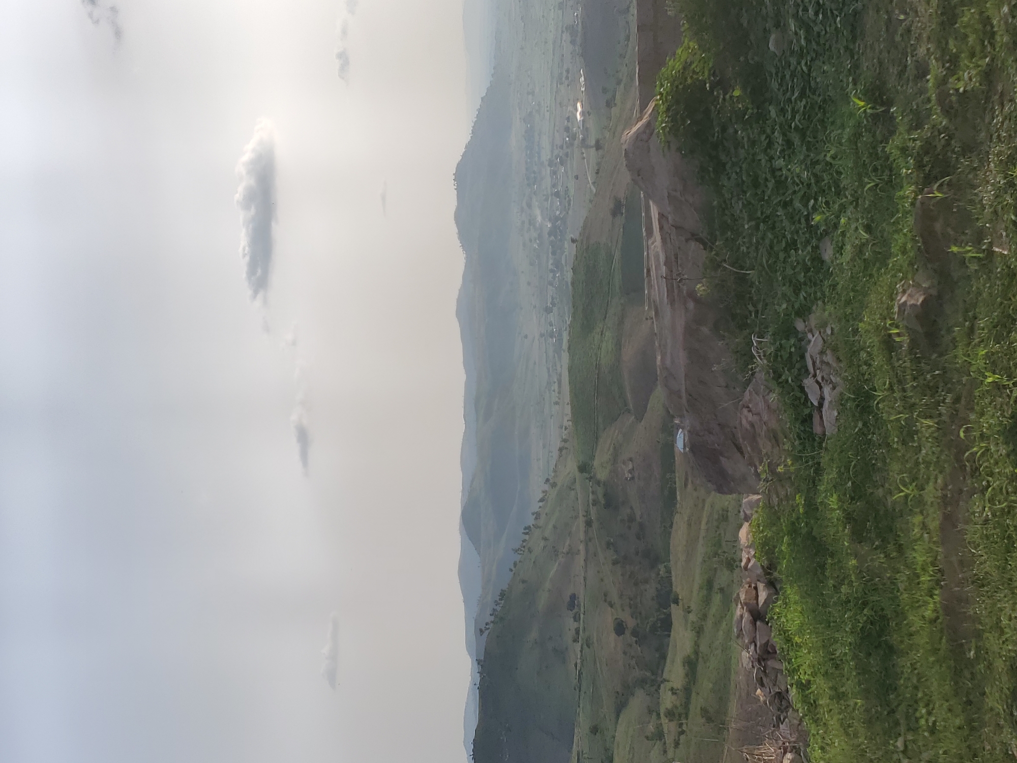 The image depicts a vast landscape dominated by rolling hills and mountains under a hazy sky with scattered clouds. The foreground features a patch of rough terrain with rocks and dense green vegetation. Further into the scene, the landscape transitions to more cultivated or naturally formed hills, with a scattering of trees and a small village or settlement visible in the valley. The overall impression is one of a rural, possibly mountainous region, with a sense of distance and depth conveyed by the layering of hills and atmospheric haze.