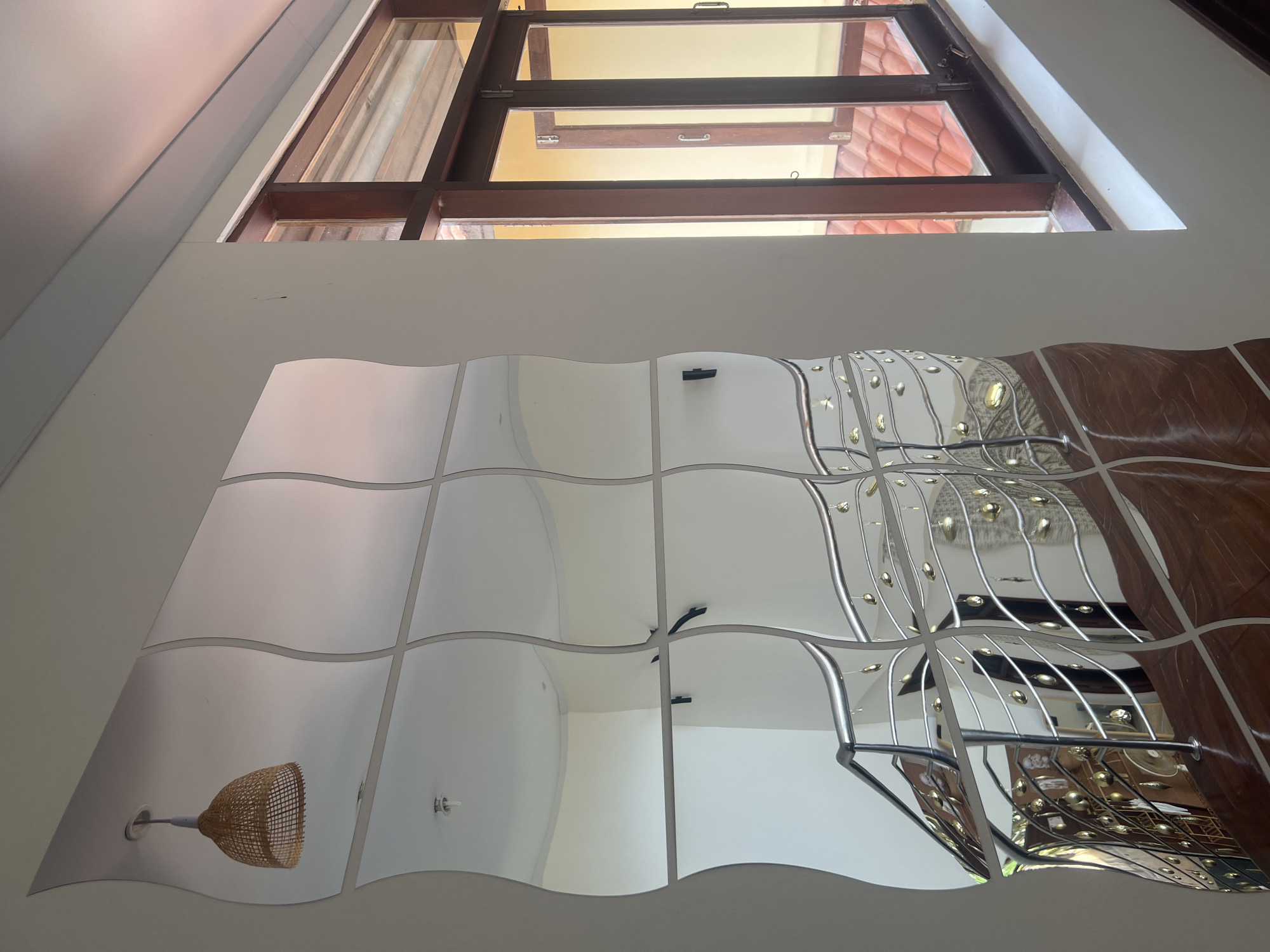 The image shows an interior scene with a wall covered in wavy mirror tiles on the left and a window with wooden frames on the right. A woven pendant lamp hangs from the ceiling above the mirror tiles. Reflected in the mirrors are a staircase with a chrome railing decorated with small gold ornaments, a ceiling fan, and a light fixture. The window looks out onto a tiled roof and a building with yellow walls. The scene appears to be a hallway or staircase area within a building, possibly a hotel or residential property, located in Ko Samui District, Thailand, as suggested by the architecture and outdoor elements visible through the window. There are no people present in the image.