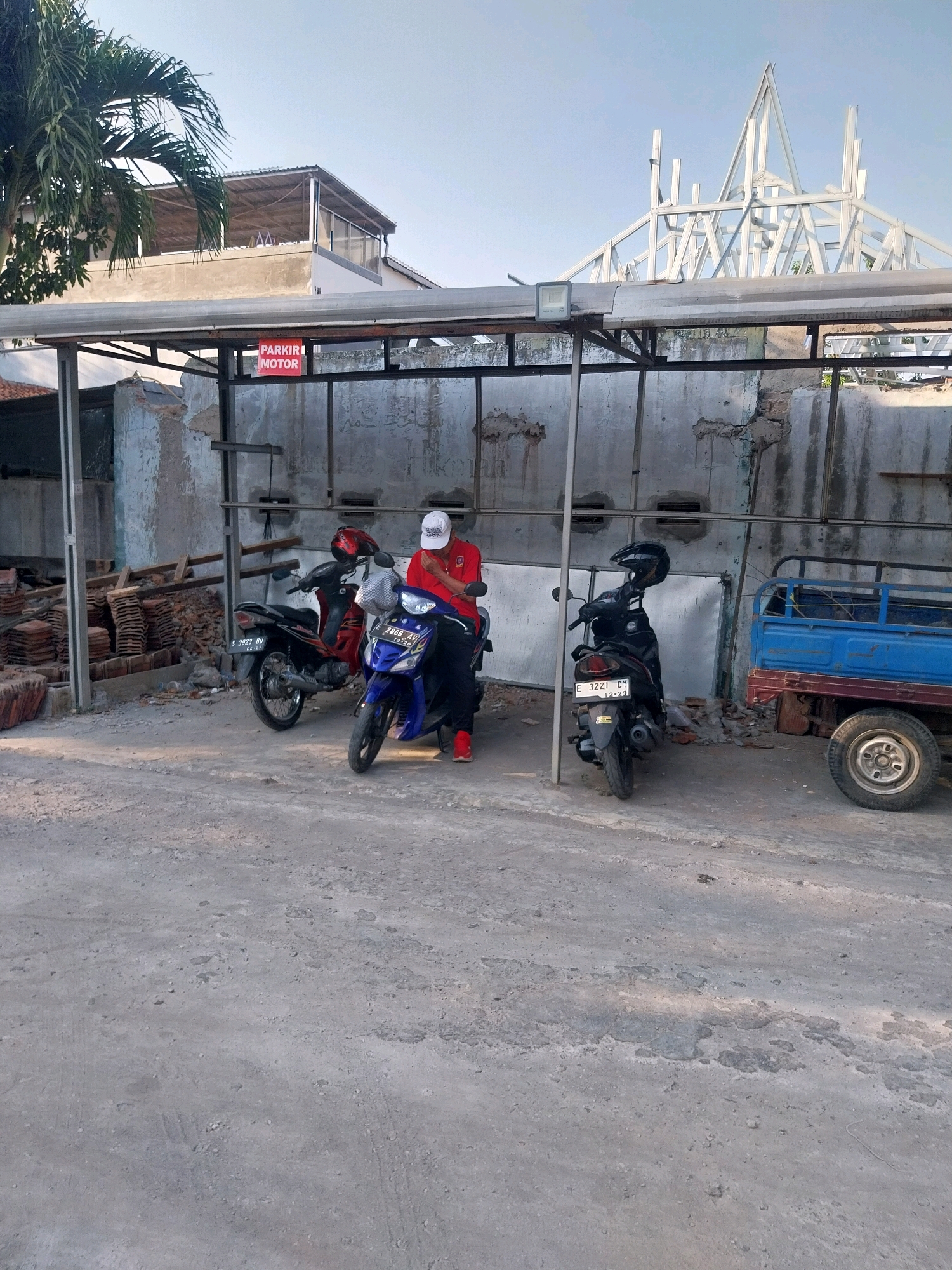 A man wearing a red shirt, white cap, and black pants stands beside a blue motorcycle under a covered parking area. The parking area has a metal roof and concrete walls. Behind the motorcycles, there is a pile of stacked bricks and a blue truck with a metal bed. In the background, a building with a metal roof is under construction. The scene appears to be in an outdoor area with a concrete ground surface. The sign "PARKIR MOTOR" is visible on a red rectangle hanging from the roof.