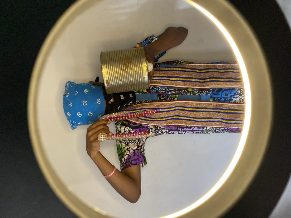 A partially visible individual stands within the circular illumination of a ring light against a plain white background, indicative of an indoor setting in Ngleshi Amanfro, Ghana. The person wears a blue bandana with white patterns covering their head, a black face mask obscuring their lower face, and a multi-colored shirt featuring purple, green, blue, and white batik-style patterns, with two vertical strips of woven fabric in brown, blue, and yellow stripes. A pink wristband is visible on the left wrist. The left hand displays a prominent gold-colored, mesh-patterned ring on the ring finger and holds multiple strands of pink and clear beaded necklaces. The right hand supports a cylindrical, ribbed metal can with a slightly tarnished, golden hue. The person's face is largely obscured.