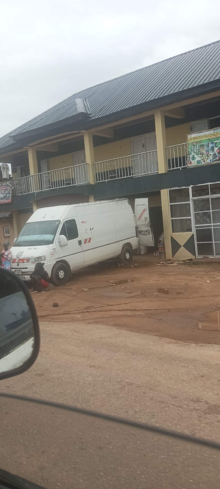 The image captures a scene in Durumi, Nigeria, featuring a white van tilted at an angle near a two-story building. A man is sitting near the front tire of the van. The surrounding ground is dirt. The building has a tin roof. The image appears to be taken from inside a vehicle, with the side mirror visible on the left.