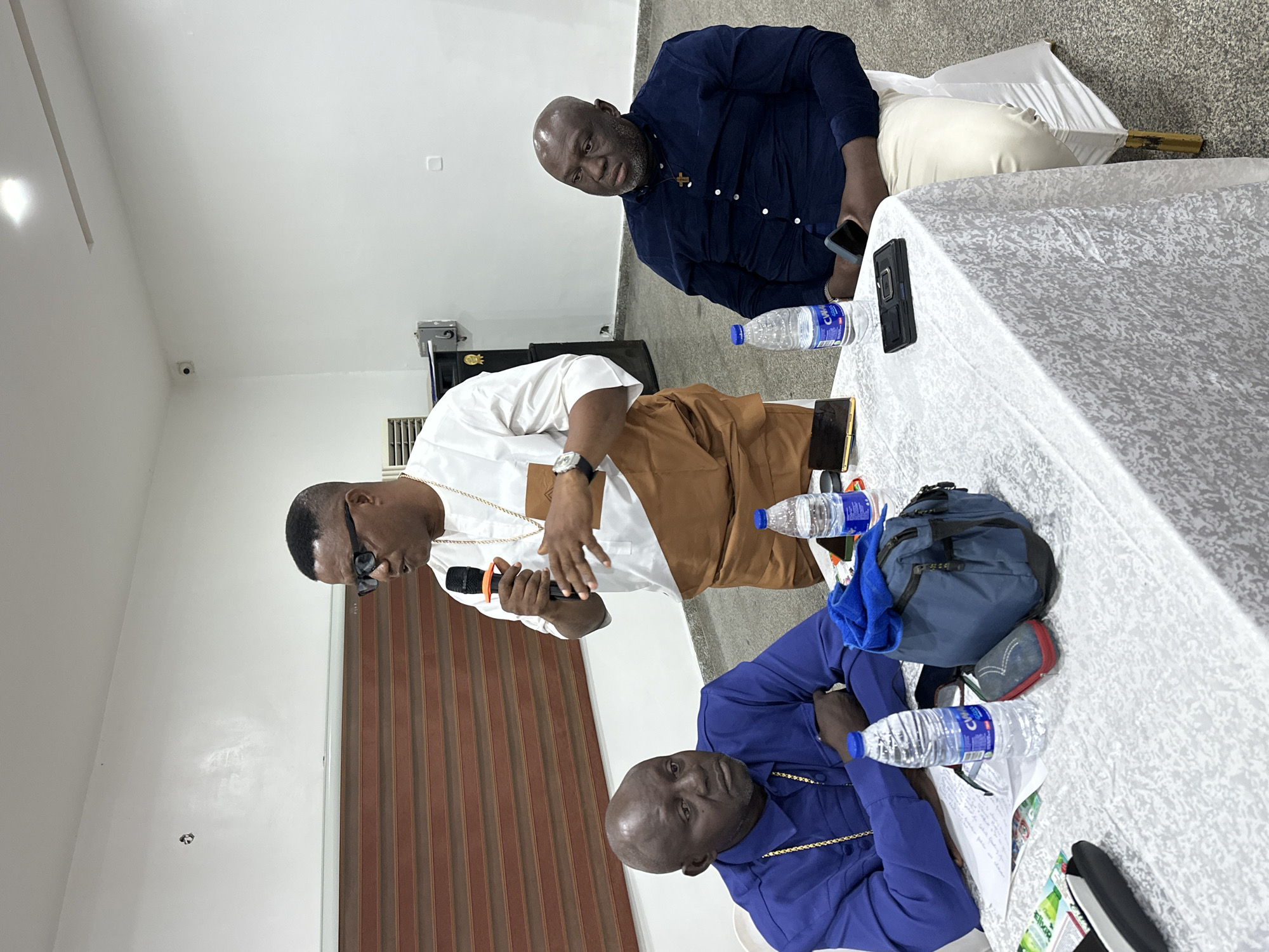 A medium shot captures three Nigerian men in a brightly lit indoor room, likely a conference or meeting space in Abuja. The room has white walls and a textured, horizontal-slatted window blind. The floor is a speckled grey. In the foreground, a white tablecloth covers a table laden with items: two bottles of water, a blue toiletry bag, a pair of glasses, a red and grey eyeglass case, and papers.
The man on the far left, seated and wearing a royal blue dashiki and a gold chain, looks towards the speaker with a serious expression, his arms crossed. To his right, the man standing, dressed in a white and brown traditional outfit and glasses, holds a microphone and gestures with his other hand, actively speaking. A smartwatch is visible on his left wrist. On the far right, another man, seated, wears a dark blue button-down shirt with a cross pendant and light-colored trousers. He looks towards the speaker, holding a dark smartphone in his lap. The overall atmosphere suggests a formal discussion or presentation.