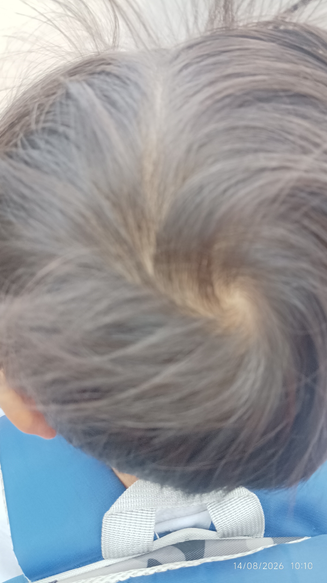 The image shows a close-up, top-down view of a person's head with dark hair, featuring a prominent swirl pattern at the crown. The hair is somewhat fine and appears to be styled or naturally falling in a circular direction. The lighting highlights the texture of the hair. The background includes a bright white area at the top and a blurred blue object with white straps and a gray pattern in the lower portion of the image, suggesting the person is wearing or sitting on something. A timestamp in the bottom right corner reads "14/08/2026 10:10". The image is a photograph taken in Tasikmalaya, Indonesia.