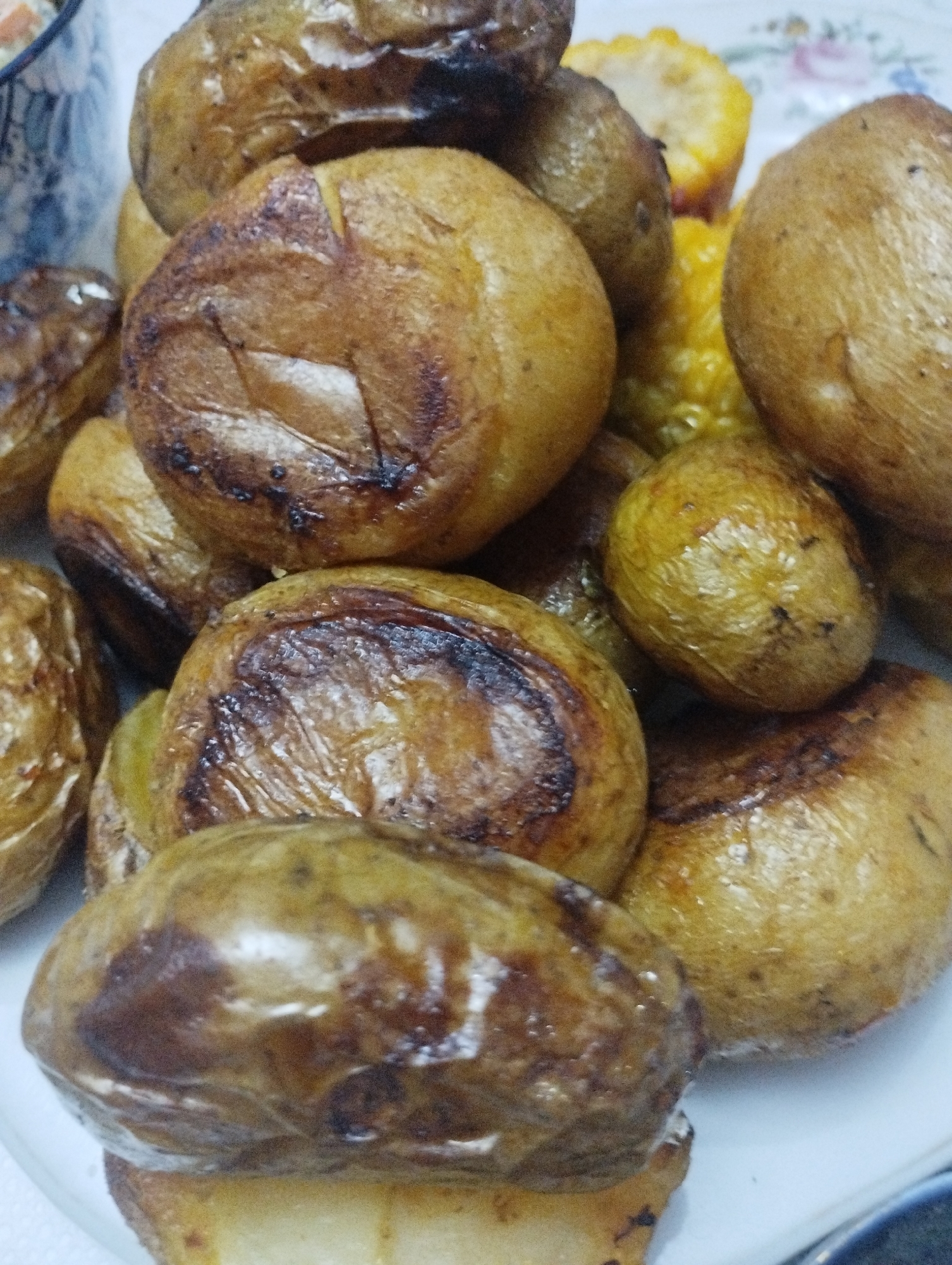 The image displays a close-up, top-down view of a pile of roasted potatoes and corn on a white plate. The potatoes are whole and vary in size, with a golden-brown, slightly charred skin. Some have visible cuts or indentations. The corn appears to be kernels, yellow in color, and mixed in with the potatoes. The lighting is bright, creating reflections on the potato skins. The background is blurred, but suggests a table setting with a hint of a patterned object and a partially visible blue and white patterned bowl or container. There are no people visible in the image, and no discernible features indicating a specific location within Bilajari, Azerbaijan. The scene is purely a still life of food.