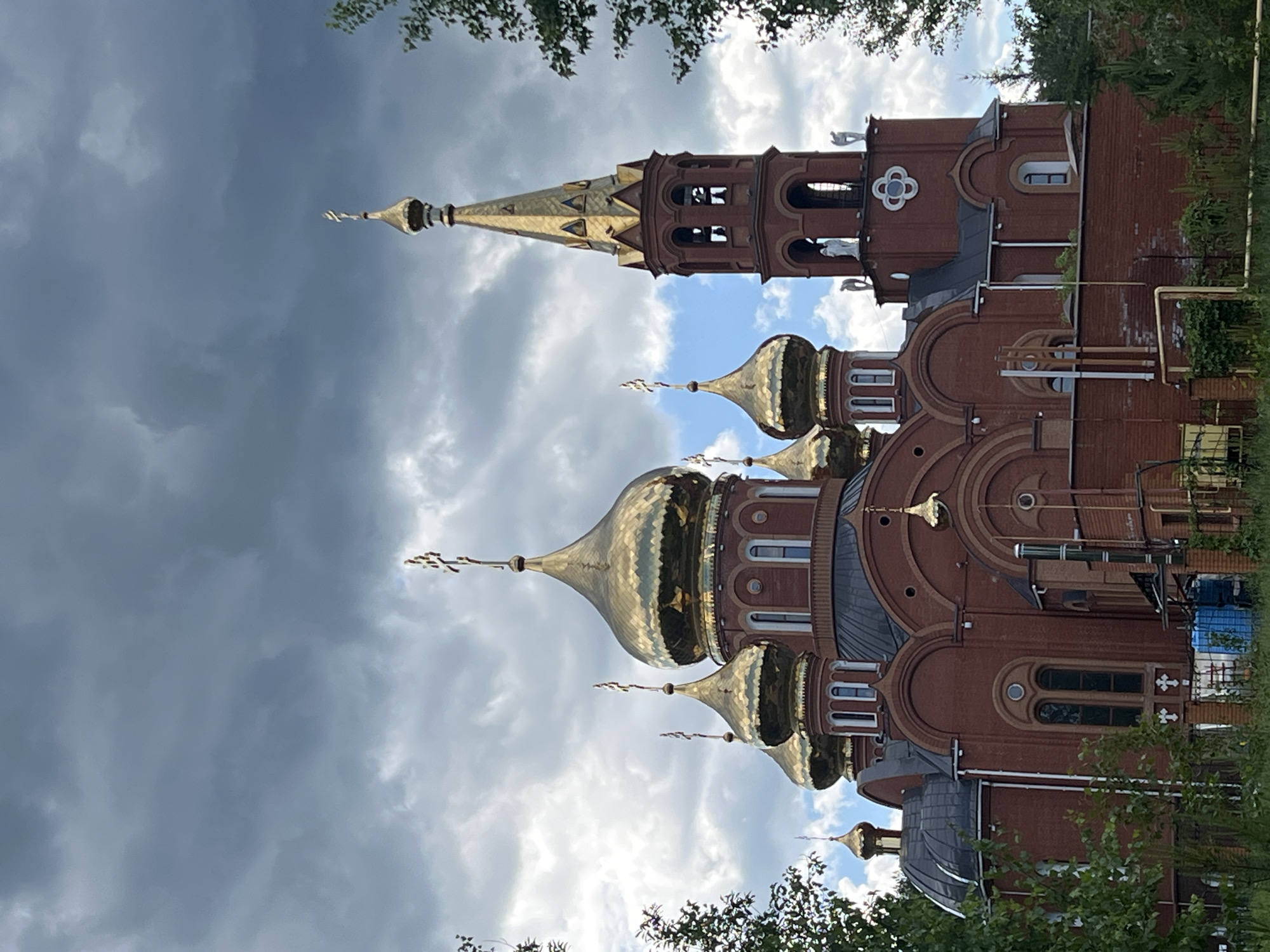 This image depicts an Orthodox church located in Chaikovsky, Russia. The central subject is a multi-domed, red brick church under an overcast sky.
The church features prominent golden onion domes, at least five of which are large and centrally positioned on the main structure, each topped with a cross. Additional smaller golden domes with crosses are visible on subsidiary structures. The main building material is red brick, characterized by arched windows with white frames, dark gray or black sloped roofs, and rounded architectural elements. To the right of the main dome cluster, a tall, slender bell tower rises, constructed of red brick with arched openings at two levels, housing what appear to be bells. This bell tower is capped with a golden, conical spire and a cross. White statues, possibly angels, are positioned on pedestals near the top of the bell tower, below the spire. A distinctive white quatrefoil decorative element is present on the bell tower's lower facade.
The immediate foreground includes dense green foliage, comprising trees, shrubs, and grass, partially obscuring the lower sections of the church. A brick wall with a metal fence runs horizontally across the bottom of the frame. Behind this fence, on the church's lower left side, blue plastic barrels or containers and various pipes are visible against the brickwork. More green trees frame the church on both the left and right sides.
The sky is predominantly covered with dark gray and lighter gray clouds, indicating an overcast or potentially stormy weather condition. Patches of lighter sky, with hints of blue, are visible between the clouds, particularly around some of the golden domes in the upper right portion of the image. No people are visible in the scene.