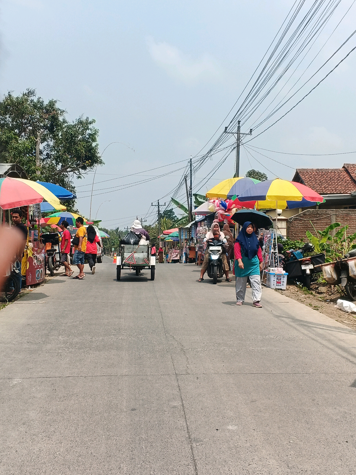 The image shows a street scene in the city of Karangjengkol, Indonesia. The street is lined with vendors and stalls, with colorful umbrellas providing shade. Several people are visible on the street. On the left, a group of people are standing near a stall. In the center of the street, a motorized cart is being pulled by a person, and on the right, a woman is walking while holding an umbrella. Further down the street, more stalls and people can be seen. The environment appears to be a typical marketplace setting on a bright, sunny day, with power lines visible overhead.