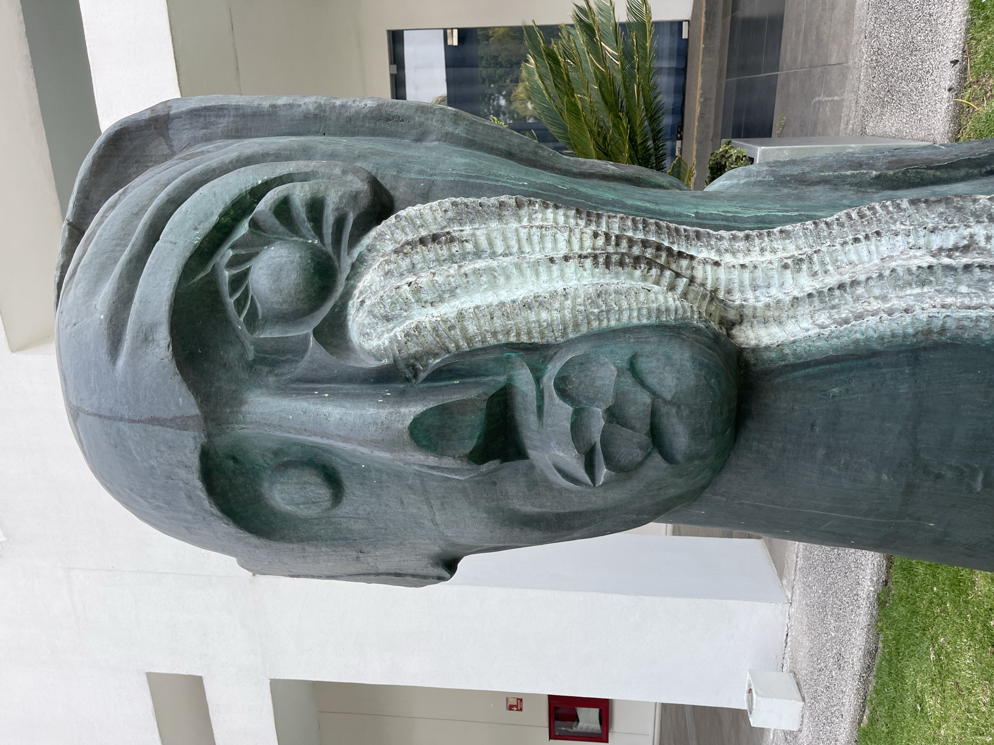 A close-up shot of a metal sculpture is set against an exterior urban background. The sculpture appears to be a head-like structure with intricate carvings and a green-gray metallic finish. The background includes a white building, a window with a partial view of interior foliage, and some greenery along the ground. The location is Unknown, Unknown.