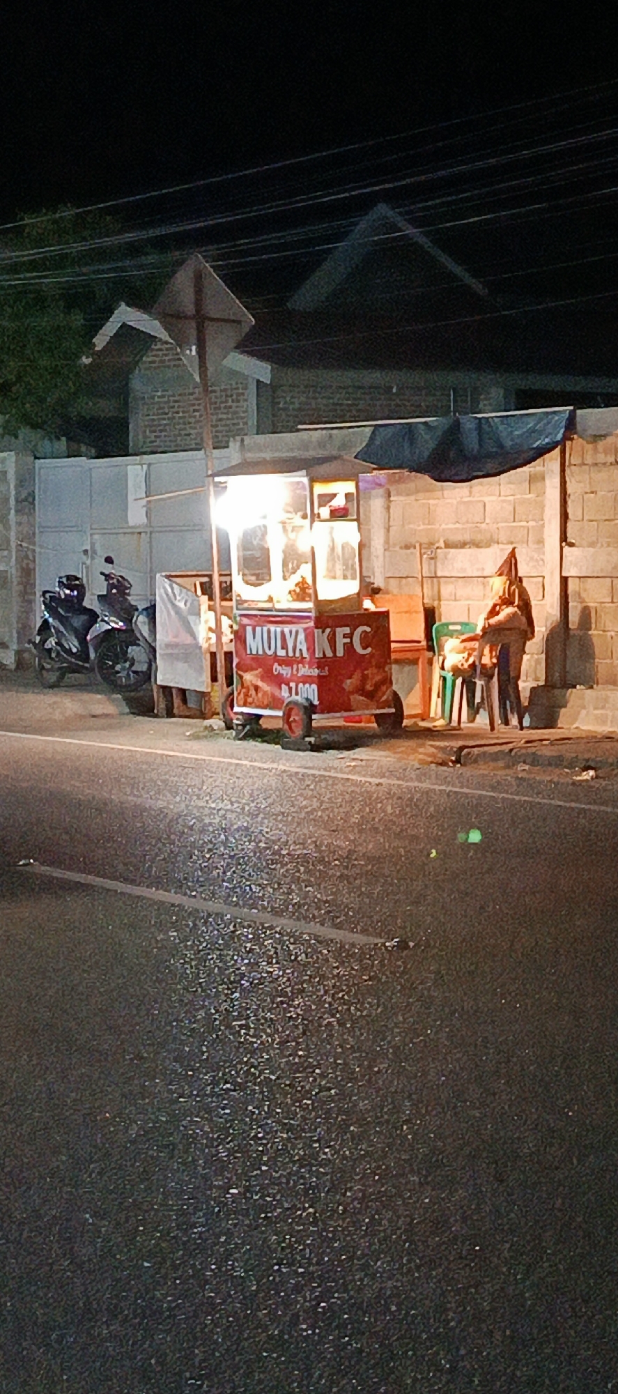In the city of Lambheu, Indonesia, at night, a food cart with the sign "MULYA KFC" is illuminated by bright lights. The cart is red and has the words "Crispy & Delicious" and a price of "7,000" visible. Behind the cart, a person wearing a head covering is seated on a green chair. Two motorcycles are parked to the left of the cart. The scene is set against a backdrop of a brick wall and a white fence. The road in the foreground is wet, reflecting the lights from the street. A traffic sign is visible on a pole near the cart.