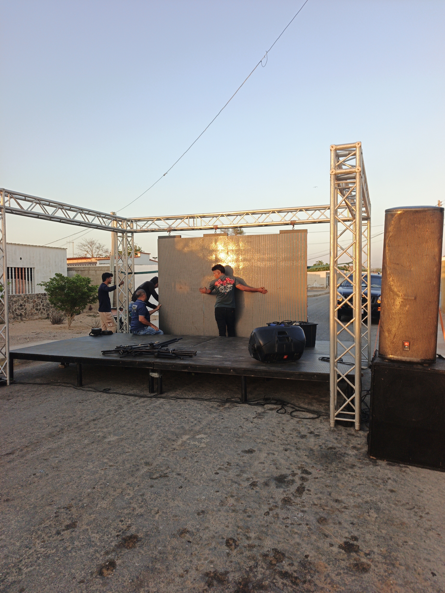 The image depicts an outdoor setting in Villa Marina, Venezuela, where a stage is being assembled. Four individuals are present on the stage, appearing to be involved in the setup process. One person is standing with their arms outstretched, seemingly aligning a large, rectangular panel, possibly part of a backdrop or display. Two others are positioned to the side, likely assisting with the panel's installation, while the fourth person is kneeling, potentially working with equipment or connections on the stage.
The stage itself is constructed with metal framework, creating a supporting structure. Various pieces of equipment, including speakers and cases, are also on the stage. In the background, a building, and a vehicle are visible, providing additional context to the location. The surrounding ground is uneven. The lighting suggests it is either early morning or late afternoon.
