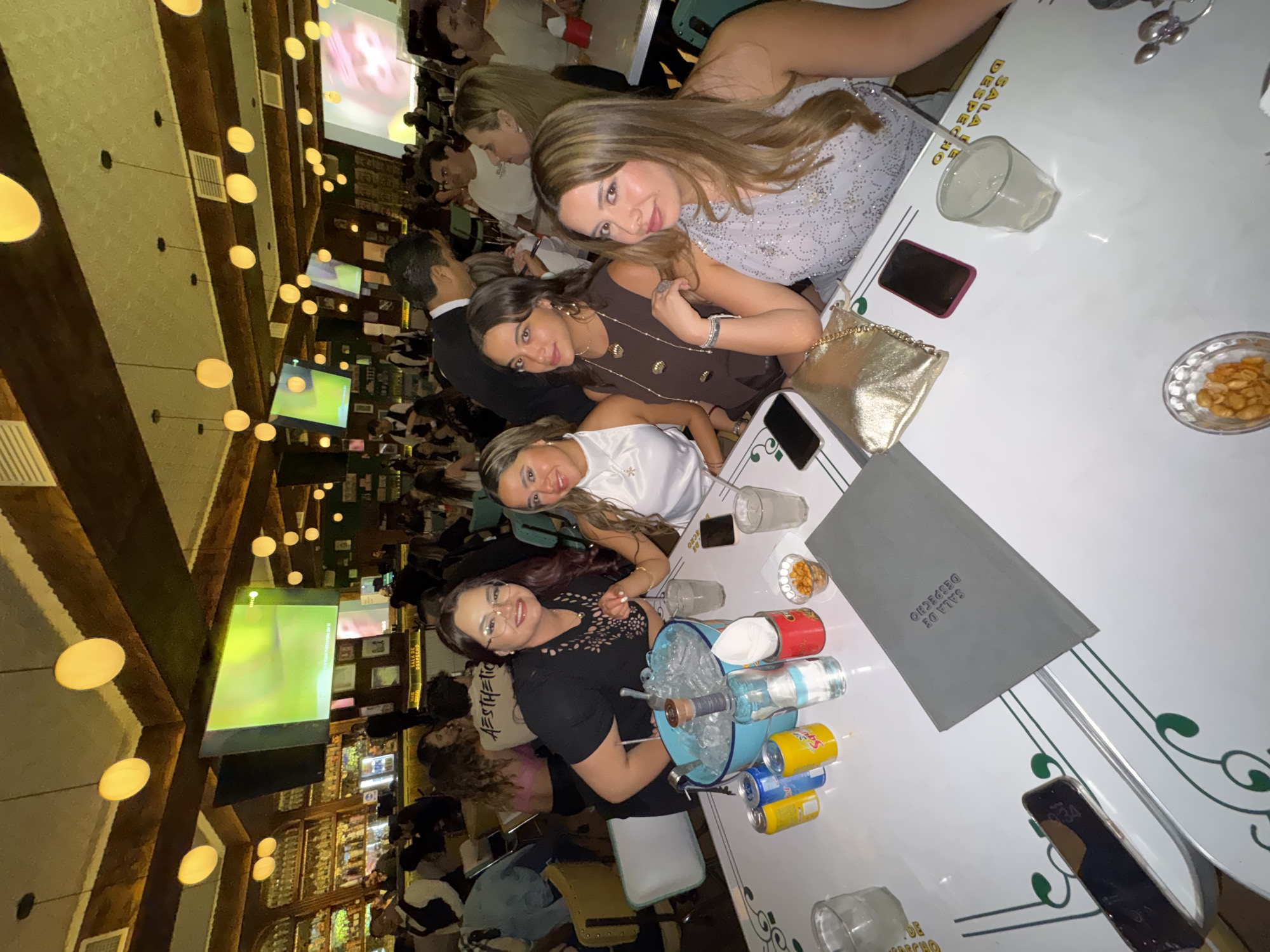 The image depicts an interior scene within a lively bar or restaurant identified as Sala de Despacho in Puebla, Mexico.
In the foreground, four adult women are seated at a white table with green decorative accents, positioned to face the camera. The woman on the far left wears a black top with cutouts and glasses, resting her arm near a light blue ice bucket filled with ice and a clear liquor bottle. Beside the bucket are three beverage cans (two yellow, one blue), a small red container with white napkins, and two clear bowls of peanuts. Two clear glasses containing drinks are also on the table, along with multiple smartphones, a gold metallic clutch bag, and a gray menu or folder embossed with "SALA DE DESPACHO". The women are smiling.
The background reveals a spacious, dimly lit establishment. The ceiling features dark wooden beams and numerous spherical, warm-toned pendant lights. Several large flat-screen televisions are mounted high on the walls, displaying various visuals. To the left, a well-stocked bar with numerous bottles and glassware is visible, staffed by individuals in vests. The entire space is populated by multiple patrons seated at various tables or standing, engaged in conversation and social interaction. Wait staff are also present, moving among the tables.