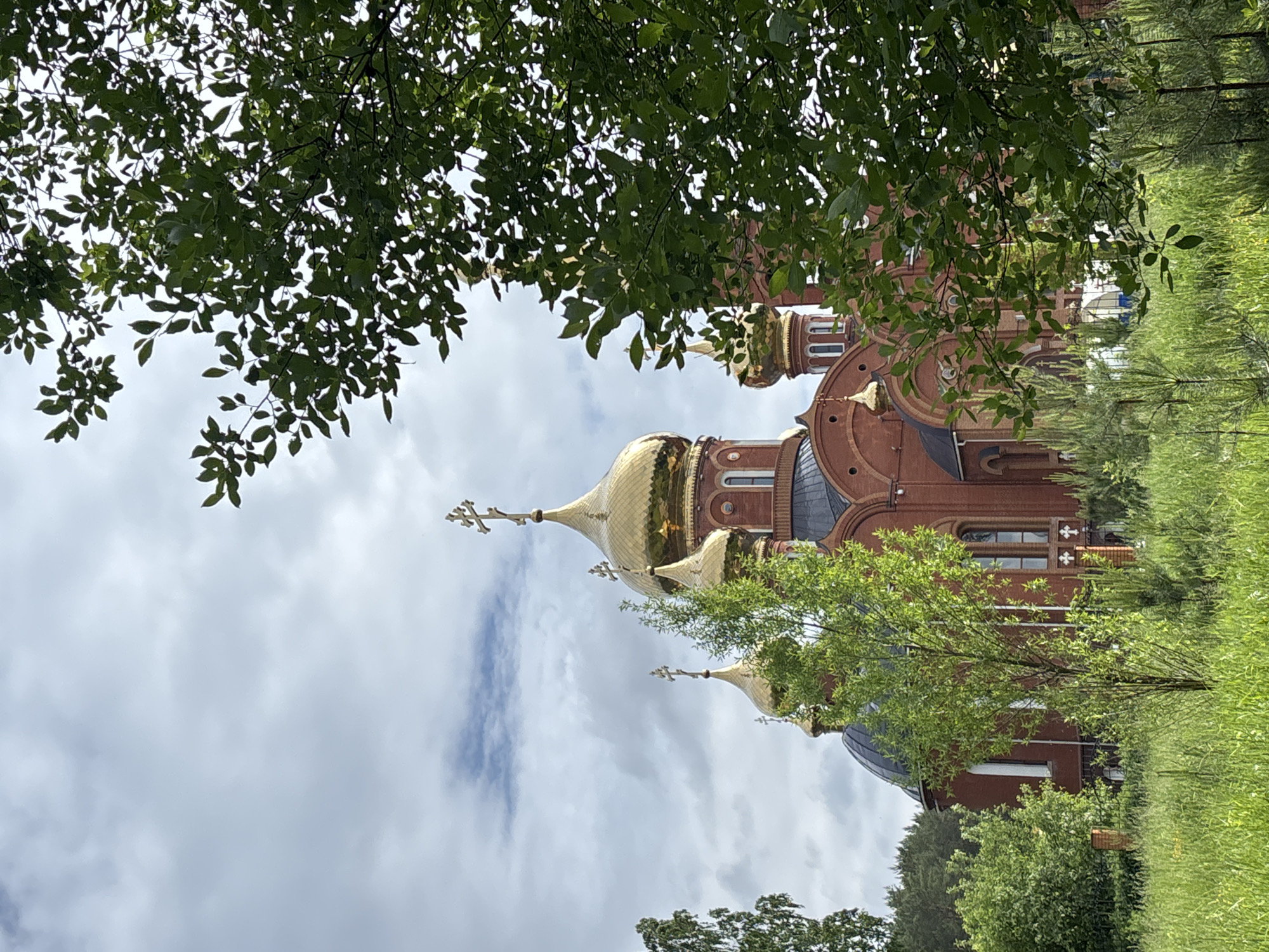 The image displays a grand Eastern Orthodox church, featuring prominent red brickwork and multiple ornate golden onion domes. The largest dome, topped with a cross, is centrally positioned, with two smaller, similarly golden domes and crosses visible to its right, all contributing to the building's distinctive silhouette. Architectural details include arched windows and intricate patterns on the domes.
Lush green tree branches, dense with leaves, partially frame the view from the top left and upper right of the composition, providing a natural foreground. The church is set within a verdant landscape, with expansive green grass and smaller trees or bushes visible around its base and to the right.
The sky is largely overcast with soft white and grey clouds, indicating daytime conditions, likely late morning or early afternoon, with diffuse natural light. No individuals or specific activities are visible, lending a tranquil and contemplative atmosphere to the scene. The architectural style strongly identifies the structure as a religious edifice, consistent with its location context in Chaikovsky, Russia. There is no visible text in the image. The overall impression is one of serene beauty and spiritual heritage nestled within a green environment.