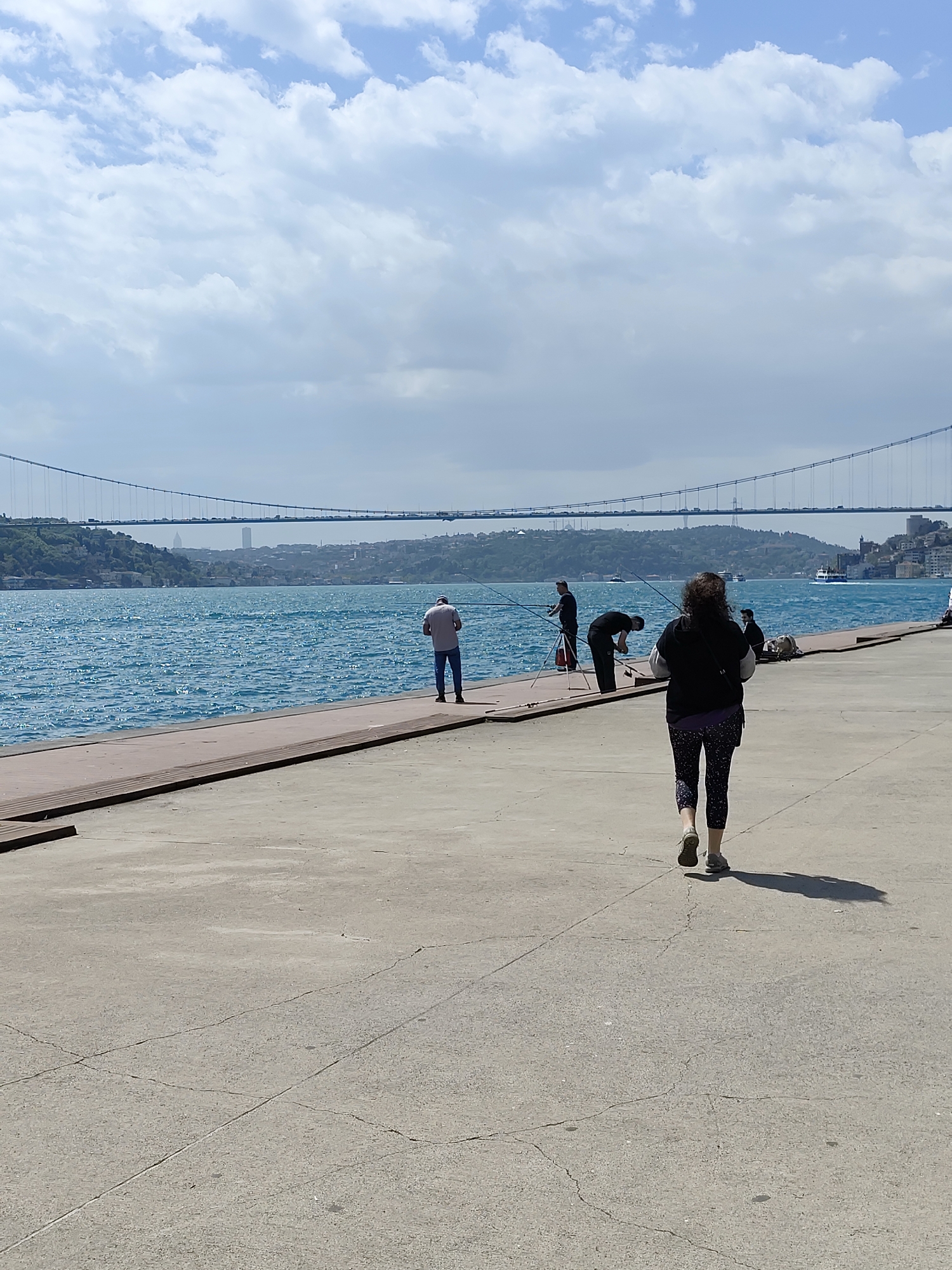 The image depicts an outdoor scene, likely a waterfront area. The environment is characterized by a concrete walkway in the foreground, leading to a lower platform made of dark wood bordering the water. Several people are present in the middle ground. Three men are engaged in fishing, each with a fishing rod. A woman in dark clothing is walking away from the viewer, facing the water. The water itself is a clear blue, with gentle waves. In the background, a suspension bridge is visible, connecting land masses on either side of the water. The sky is partly cloudy, with areas of blue visible. The location appears to be in Istanbul, Turkey, due to the presence of the Bosphorus Bridge.
