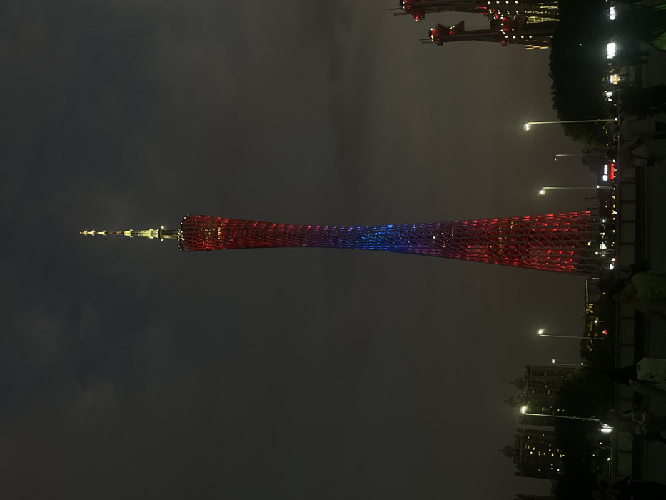 The Guangzhou Tower, also known as the Canton Tower, is lit up in red and blue lights, in the city of Guangzhou Shi, China, at night. The coordinates for the tower are latitude: 23.117, longitude: 113.319. The tower appears to be a popular tourist destination, as numerous people and streetlights are visible in the foreground. There are some building structures obscured by foliage in the bottom right corner of the image, which are likely residential. The sky is mostly dark, with a few clouds visible in the distance. The tower is the main focal point of the image, with its intricate lattice structure and illuminated colors making it the most prominent feature of the scene.