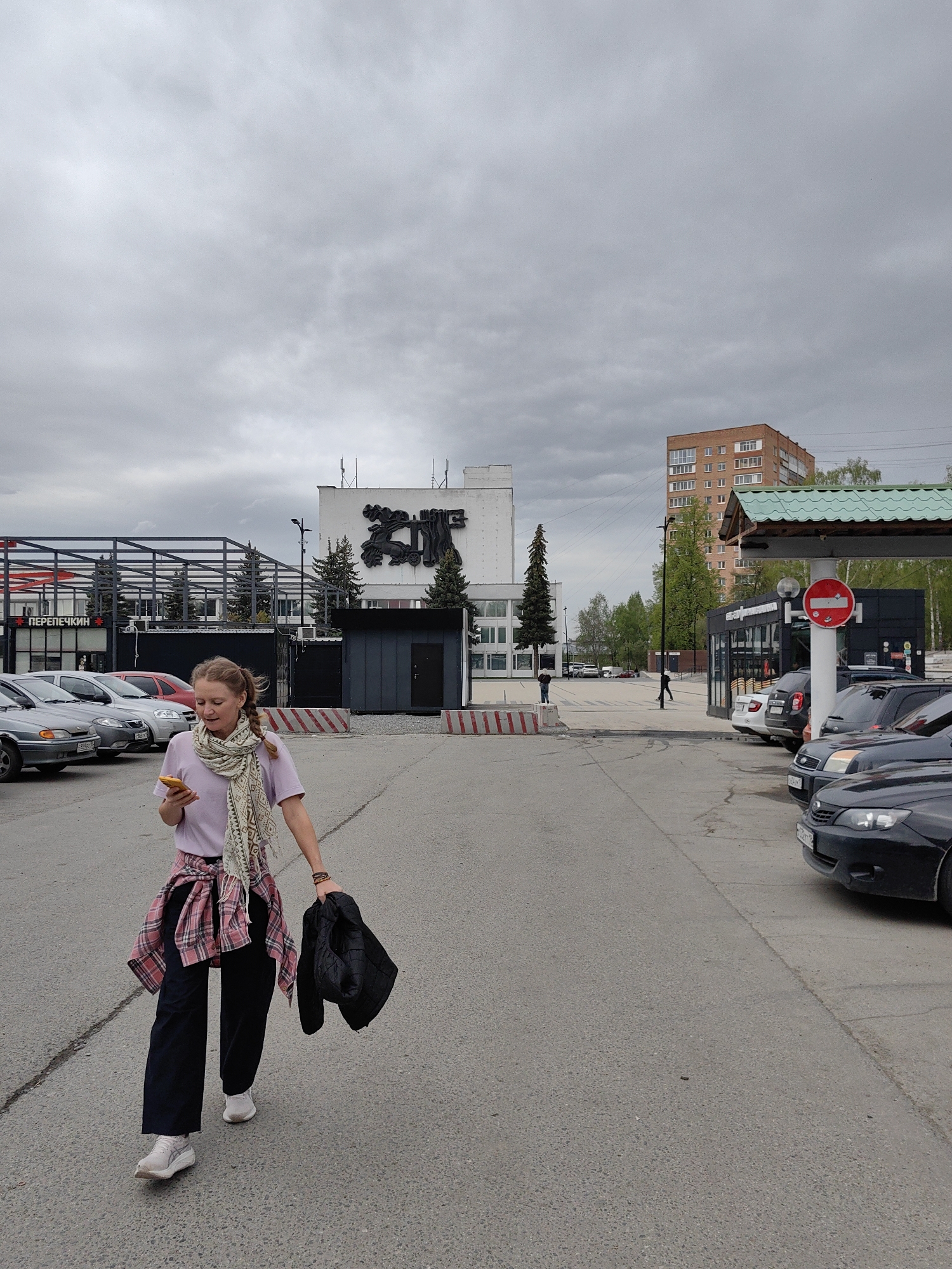 A woman walks toward the camera in a parking lot. She is fair-skinned with blonde hair in a braid. She wears a lilac-colored shirt, black pants, white shoes, and a scarf. A plaid shirt is tied around her waist, and she holds a black jacket in her left hand while looking at a yellow smartphone in her right.
Behind her, a parking lot contains several cars. In the background, there is a white building with a large black metal sculpture above its entrance and an adjacent building with a storefront. A sign above the storefront reads "ПЕРЕПЕЧКИН" in Cyrillic. Further back is a brown brick apartment building. The sky is overcast.
The scene is in Izhevsk, Russia. The white building with the sculpture is the "Rodina" cinema.