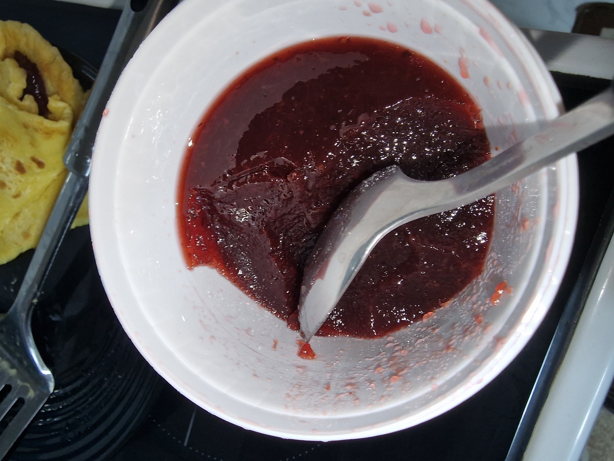 The image shows a white plastic bowl containing a dark red, possibly homemade, strawberry jam or preserve. A stainless steel spoon is partially submerged in the jam, suggesting it is being stirred or prepared for use. To the left of the bowl, a folded crepe or pancake with a filling similar in color to the jam is visible on a black surface, likely a stovetop. The setting is indoors, possibly a kitchen environment.