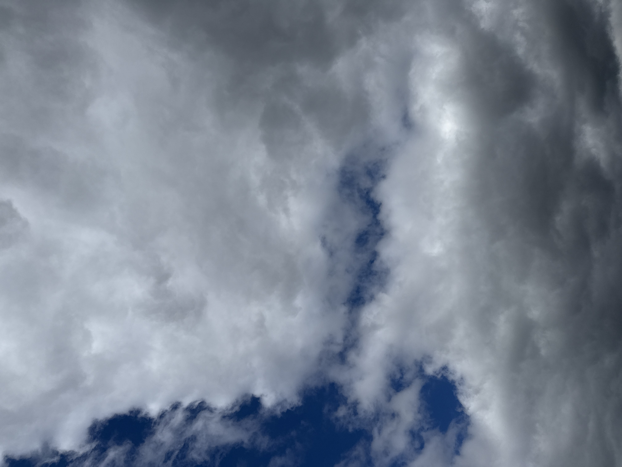 The image is a close-up, low-angle shot of a cloudy sky. The sky is a mix of dark gray, lighter gray, and some patches of bright blue. The clouds are textured and appear to be moving, with some areas being more solid and others more wispy. There is no indication of a specific location within Großpösna, Germany, as the image exclusively captures the sky. No people, subjects, objects, or actions are visible in the frame. The environment depicted is purely atmospheric.