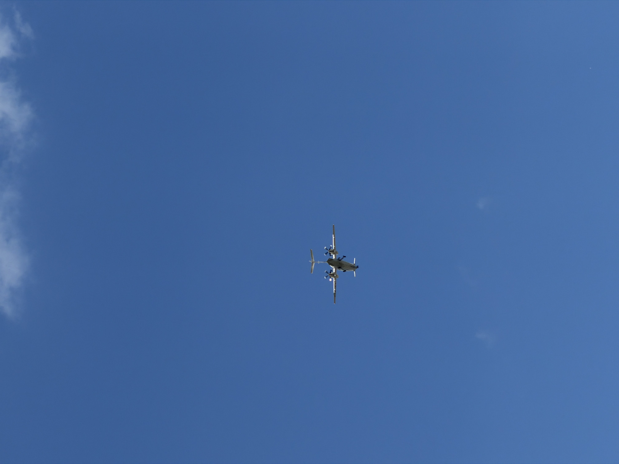 The image shows a blue sky with a small, white airplane flying in the distance. There is a wispy cloud in the upper right corner of the image. The airplane is a twin-engine propeller aircraft with its landing gear down, suggesting it is either approaching for landing or has just taken off. The scene is set against a clear blue sky. No specific landmarks or recognizable features of Burgas, Bulgaria, are visible in this image.