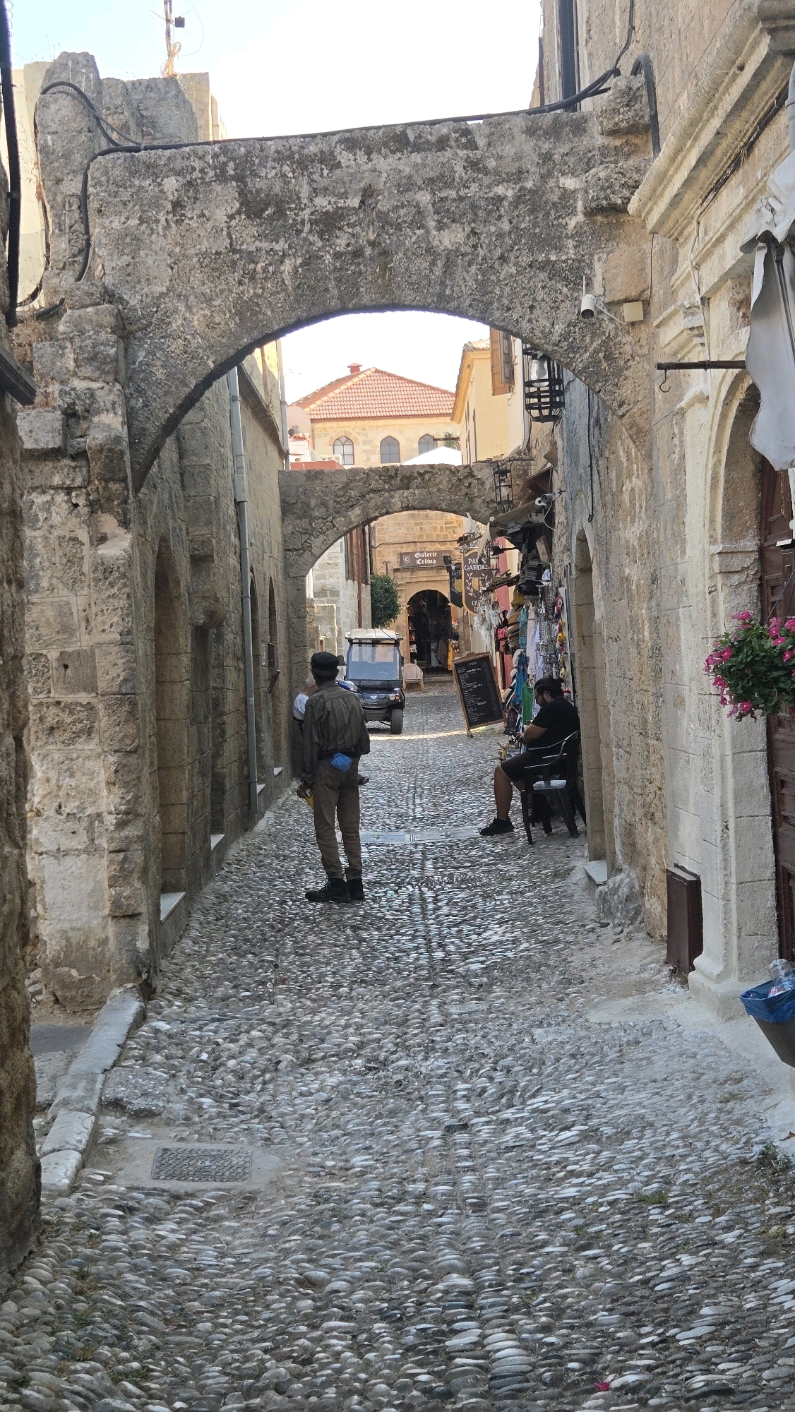 The image depicts a narrow, cobblestone street in the Municipal Unit of Rhodes, Greece, characterized by ancient stone architecture. The street is flanked by weathered stone buildings with arched doorways and windows. Sunlight casts shadows on the uneven cobblestone surface.
In the foreground, a person stands facing away from the camera, wearing a light-colored shirt, brown pants, and a black cap. They have a blue object on their lower back. To the right, a man sits on a dark chair, also facing away, wearing a black t-shirt and shorts. He is looking down.
Further down the street, a small utility vehicle is parked. Shops with various goods are visible on the right side, with awnings and displays. A black chalkboard menu stands on an easel in the street. Flowers are visible in a pot on the right. The overall scene suggests a historic, possibly tourist-oriented area.