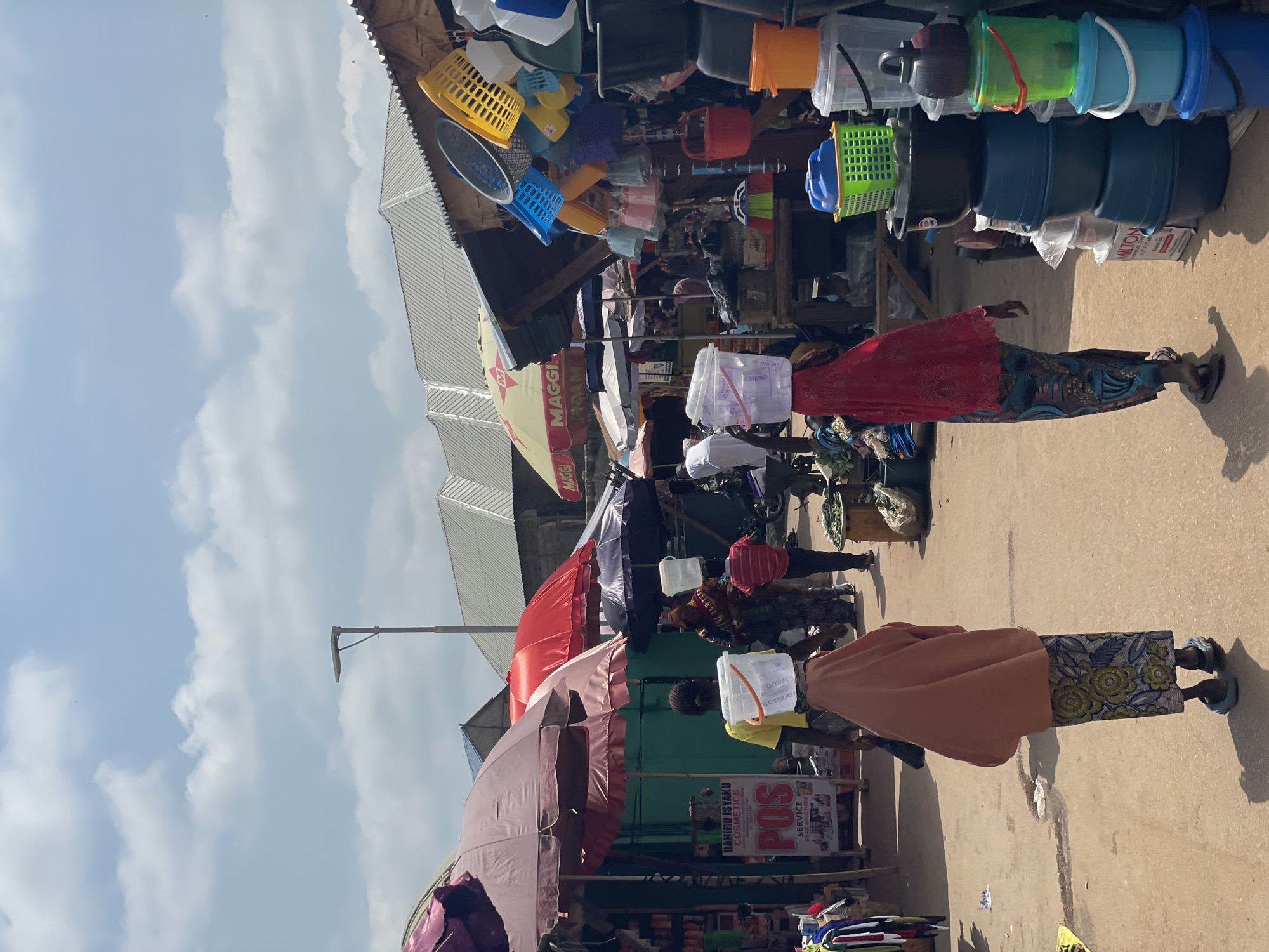 This image captures a lively outdoor market scene in Akwanga, Nigeria. The primary subjects are several individuals, predominantly women, engaged in activities typical of a marketplace. One woman, wearing a red and blue patterned wrapper and a red top, stands with her back to the camera, facing a stall filled with various plastic containers and baskets. Another woman, in a brown wrapper and a white head covering, is seen walking away from the camera, carrying a white plastic container on her head. Further back, another woman in a yellow top and patterned wrapper also carries a white container on her head, while a third person, likely a vendor, is partially visible behind a stall.
The market is crowded with a diverse array of objects, including brightly colored plastic buckets, basins, baskets, and containers stacked on shelves and hung from overhead structures. Umbrellas, in various shades of red, purple, and black, are deployed to provide shade, suggesting a sunny day. A prominent yellow umbrella with the "MAGGI" logo is visible on the left side of the frame. Textual elements, such as "MAGGI," "POS SERVICE," and "DAHIRU ISYAKU COSMETICS," are discernible on signs and packaging, indicating commercial activity. The ground is paved with concrete, and the overall setting appears to be a bustling street market under a bright sky with scattered clouds. The scene suggests a typical day of commerce and daily life in this Nigerian town.